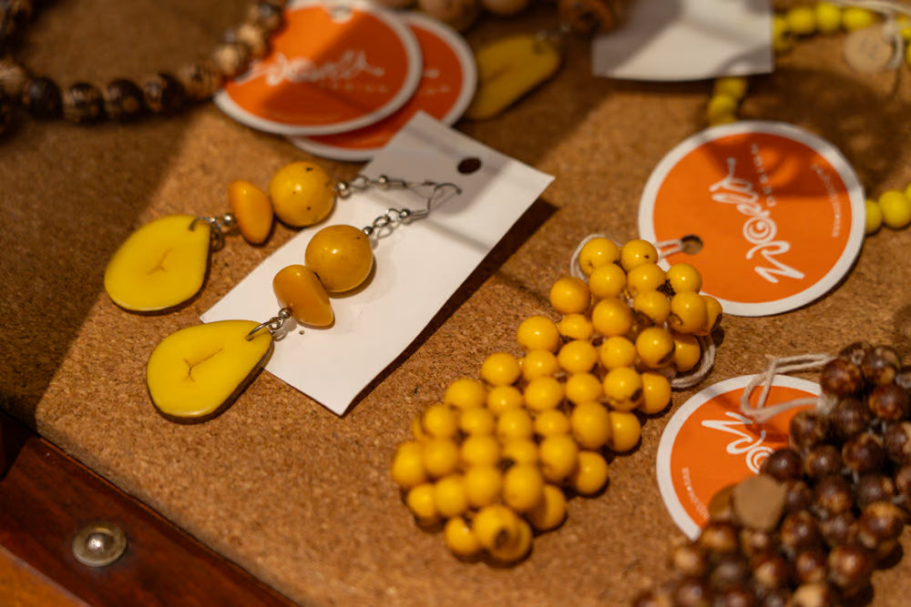 Yellow and orange beaded jewelry displayed on a cork board, including a pair of drop earrings on a white card, a cluster of round yellow beads, and other beaded necklaces with orange and white brand tags.