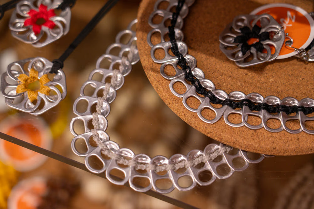 Silver bottle-cap chain necklace and matching pendant displayed on a cork stand, alongside a black cord bracelet and flower-shaped pendants.