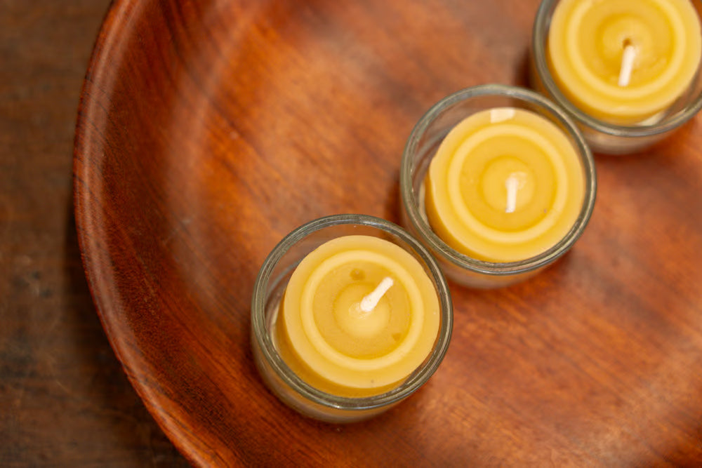 Three small yellow beeswax tea light candles in clear glass holders, arranged diagonally on a round wooden plate over a dark wood surface.