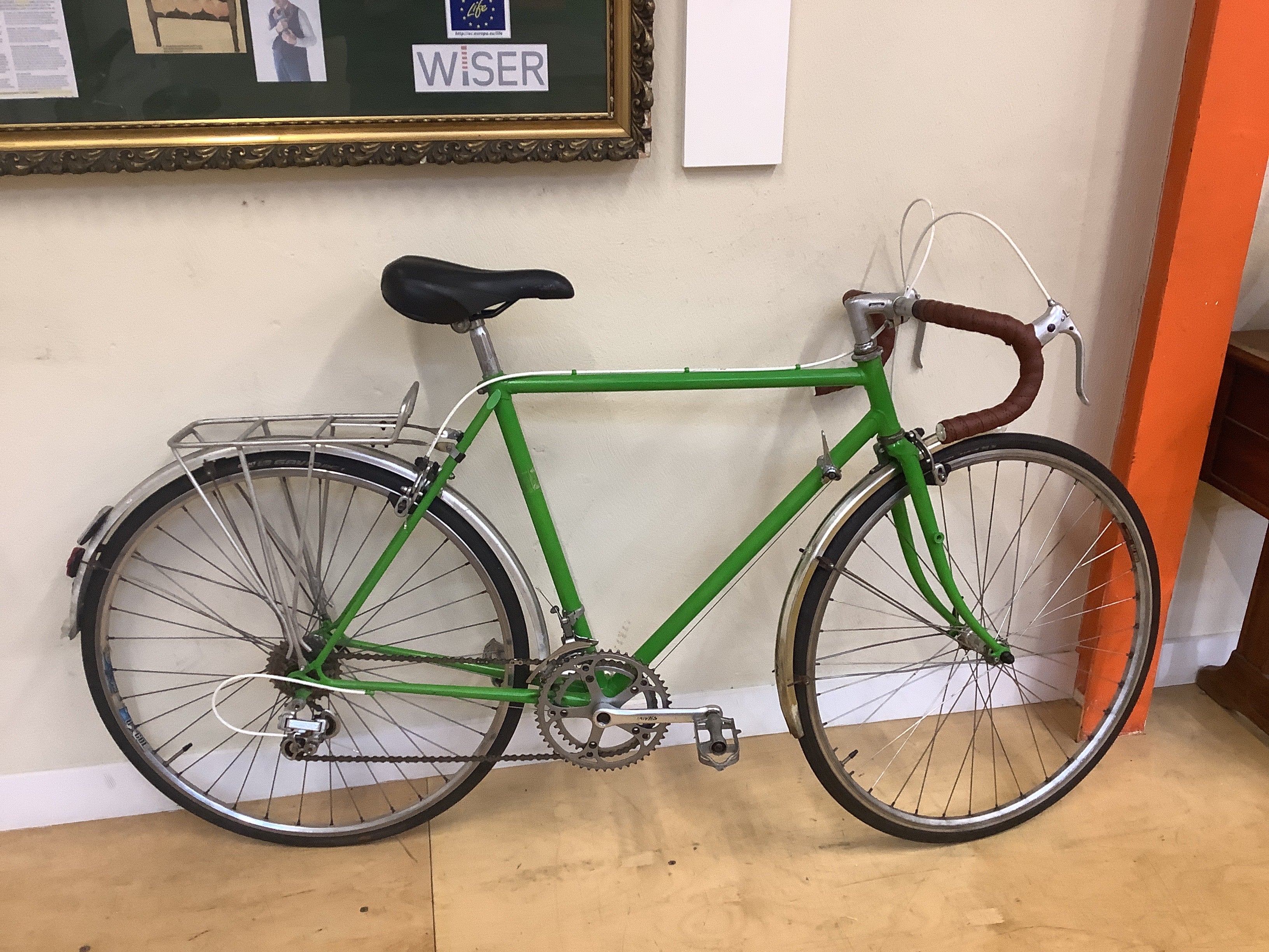 Green bicycle with brown handlebars indoors against a wall with framed pictures.