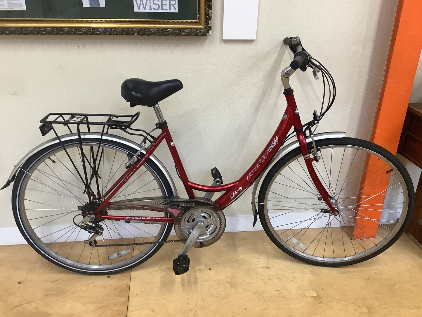 Red bicycle with a black saddle on a wooden floor against a white wall.