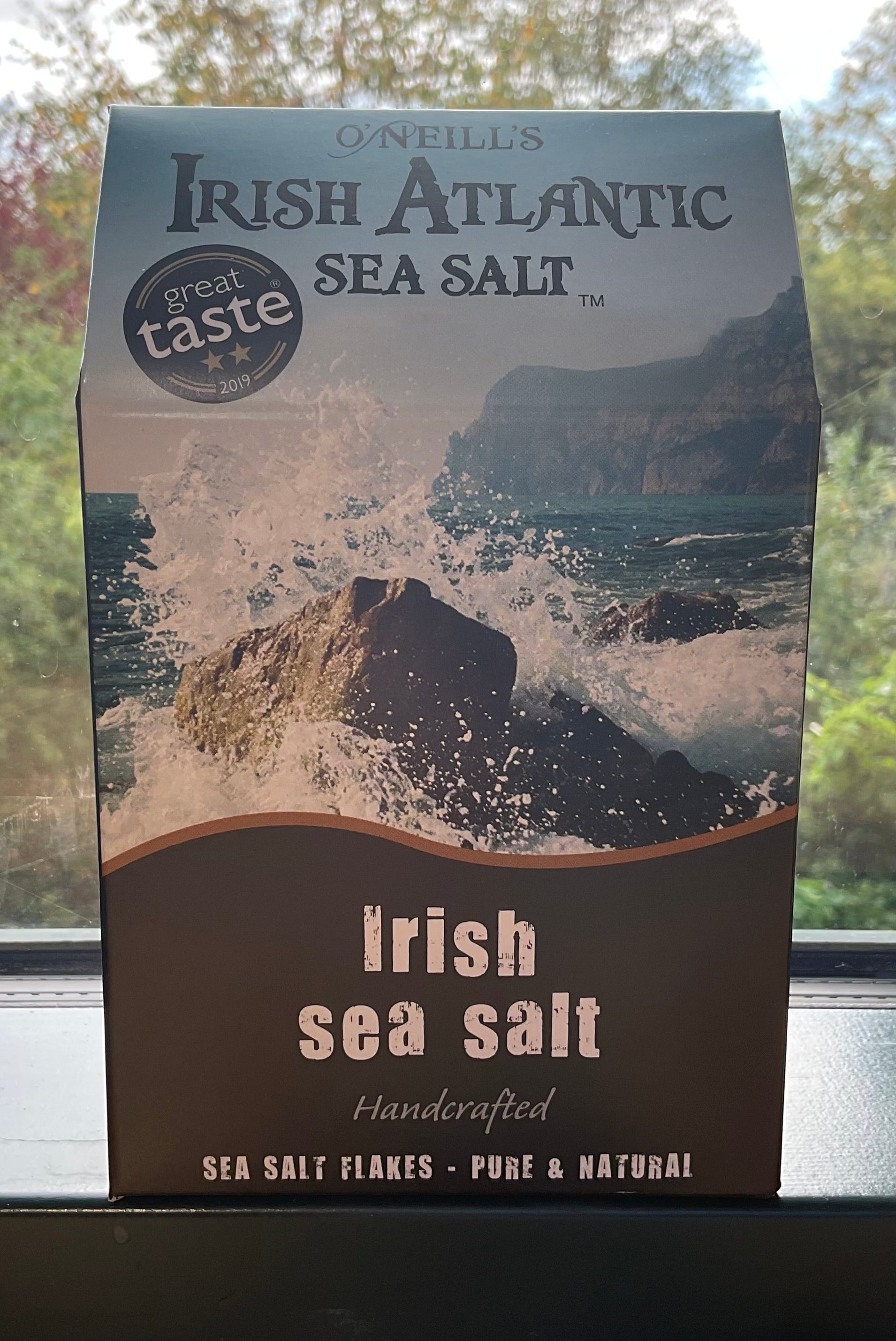 Packaging of Irish Atlantic Sea Salt Flakes with a background image of a wave crashing against rocks. The package is labeled as 'Irish sea salt' and mentions that it is handcrafted, pure & natural.