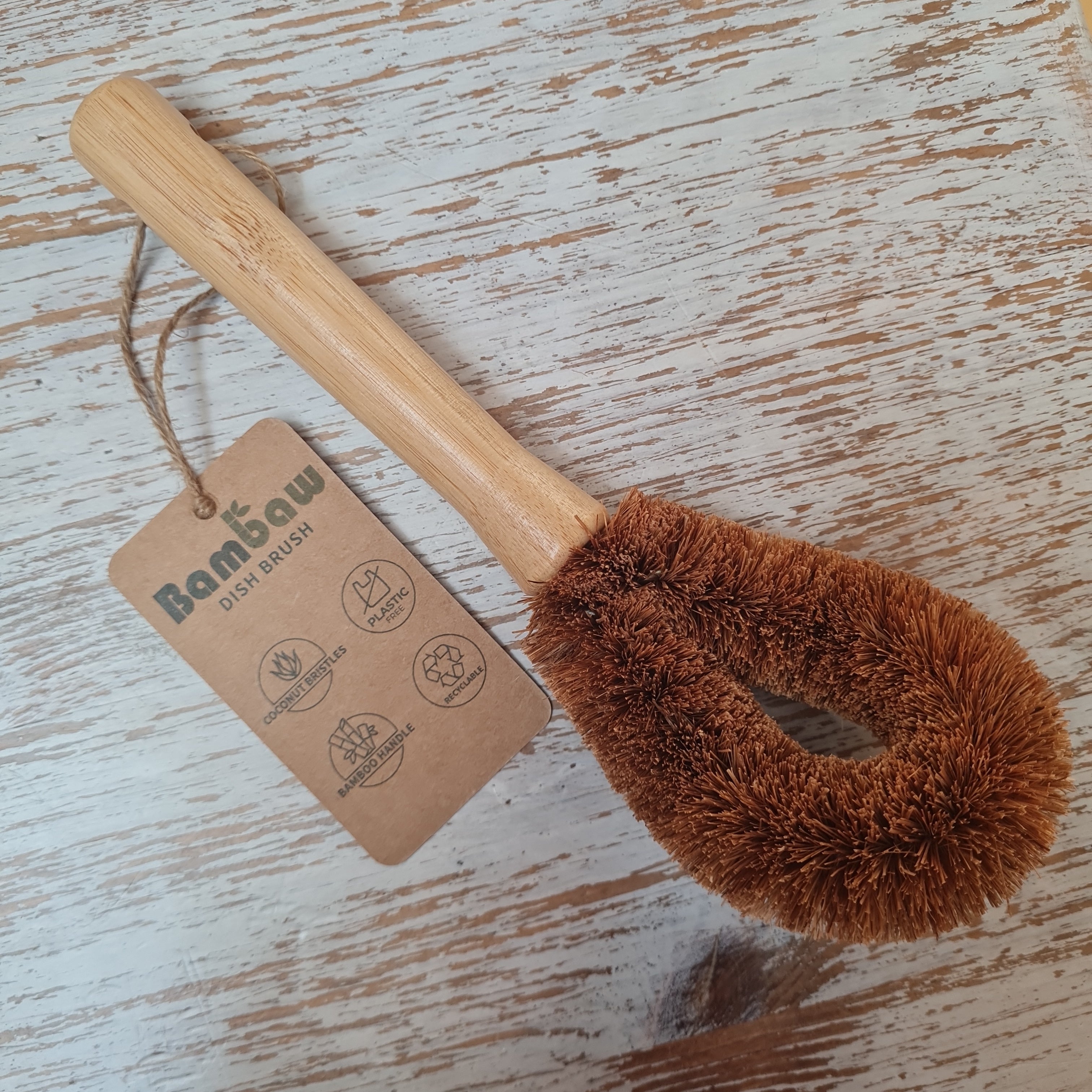 A beige dish brush with coconut bristles and a wooden handle, featuring a hanging tag with 'BAMBAW' branding.