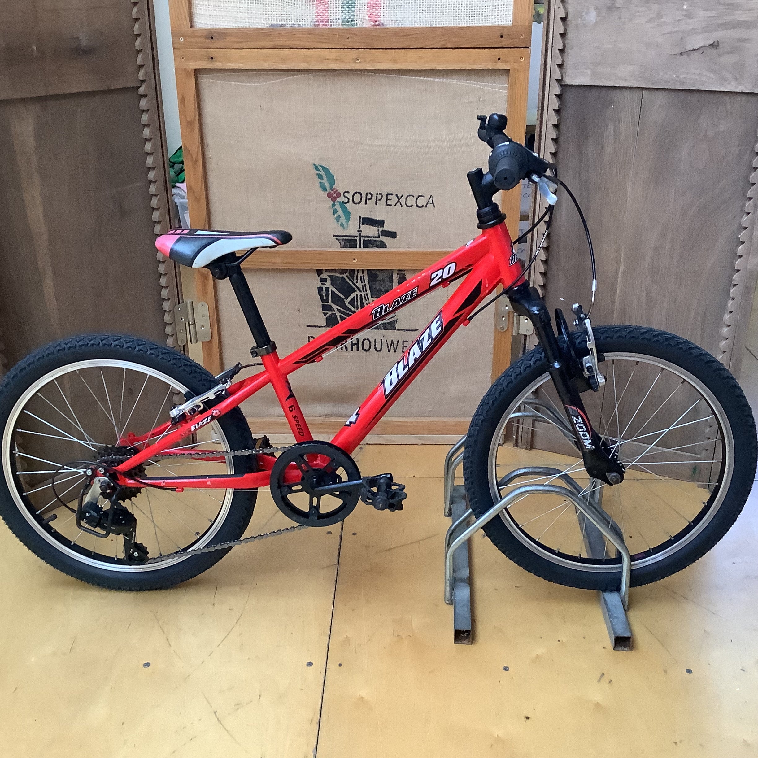 A red children's mountain bike with black tires and handlebars, displayed on a metal stand in front of a wooden door and burlap backdrop.