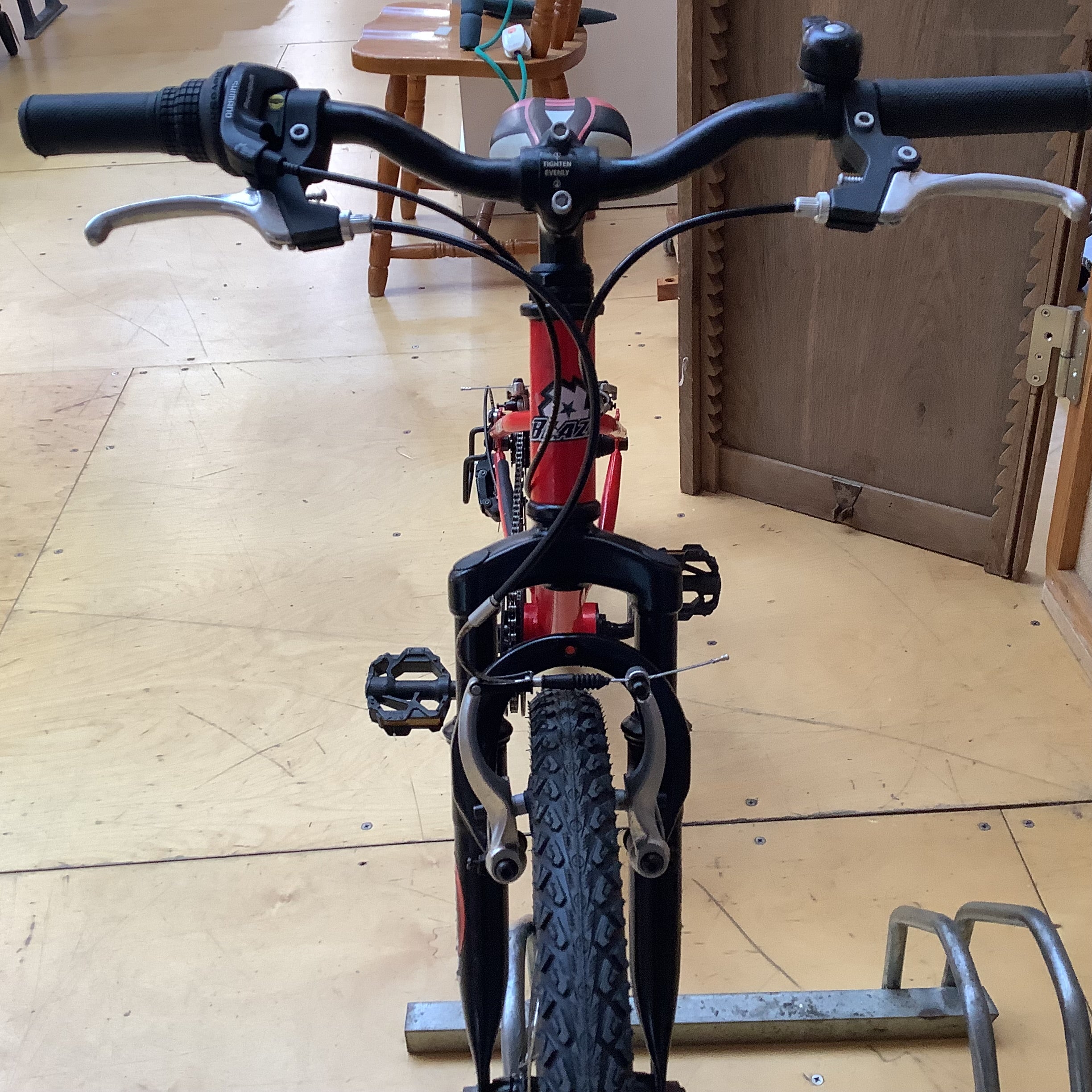 A red children's mountain bike viewed from the front, showing black handlebars, black front suspension, and a black tire, photographed indoors on a wooden floor.