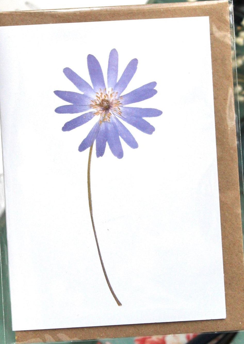 Card with a purple flower design on a white background, placed on a green surface with books and flowers.