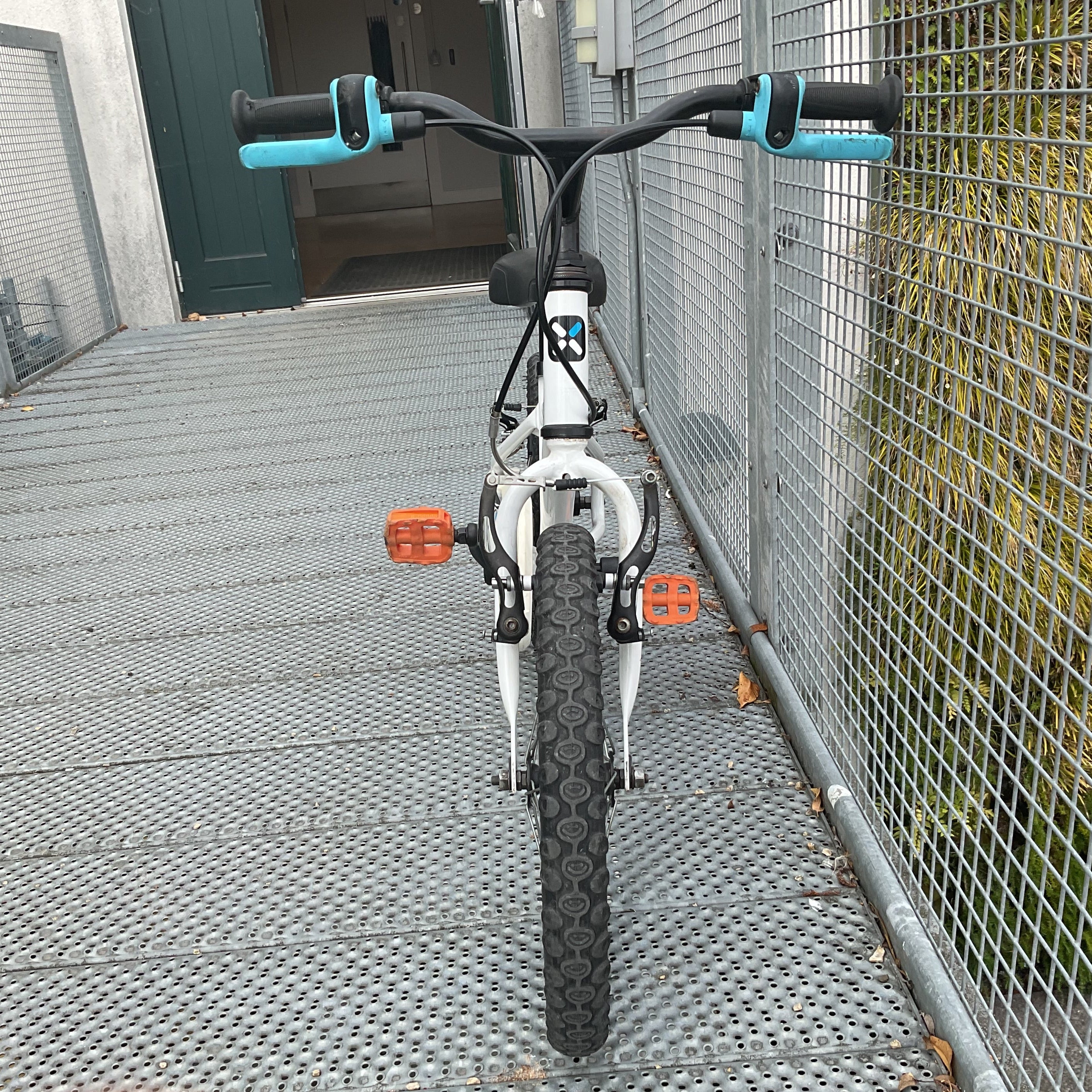 A wchildren's bicycle with a white frame, black tires, and turquoise handlebar grips, photographed from the front on a metal grate walkway next to a wire fence.