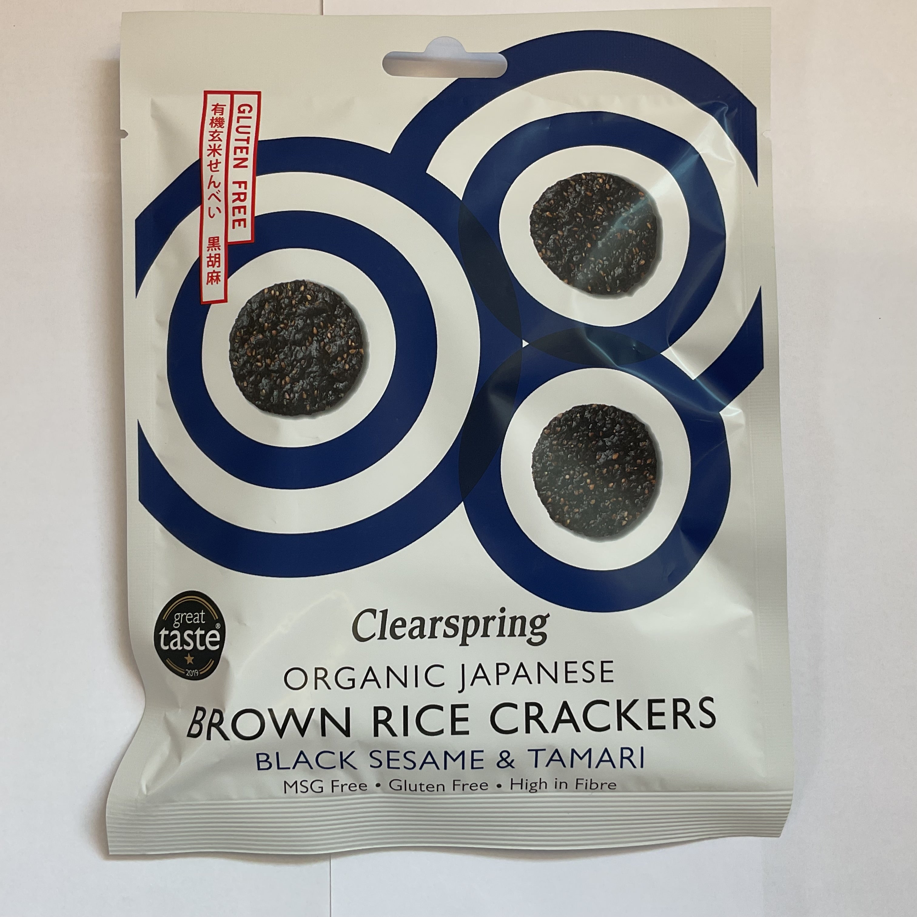 A package of Clearspring Organic Japanese Brown Rice Crackers with Black Sesame & Tamari. The packaging is predominantly white with blue and black accents, and an image of the crackers visible through a cut-out window.