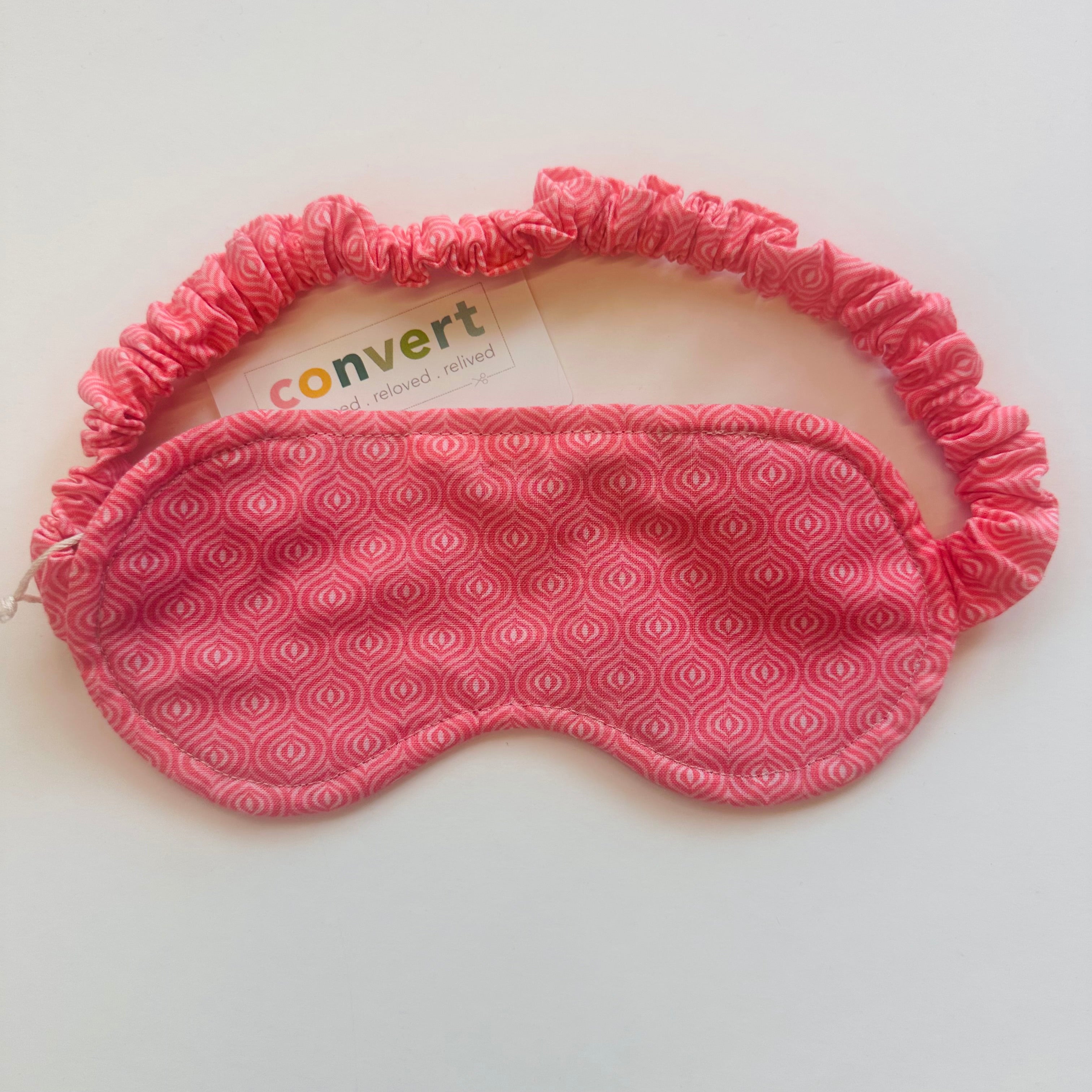 A pink patterned sleep mask with an elasticized headband, shown on a plain light background. A small card with the brand name 'convert' is visible behind the mask.