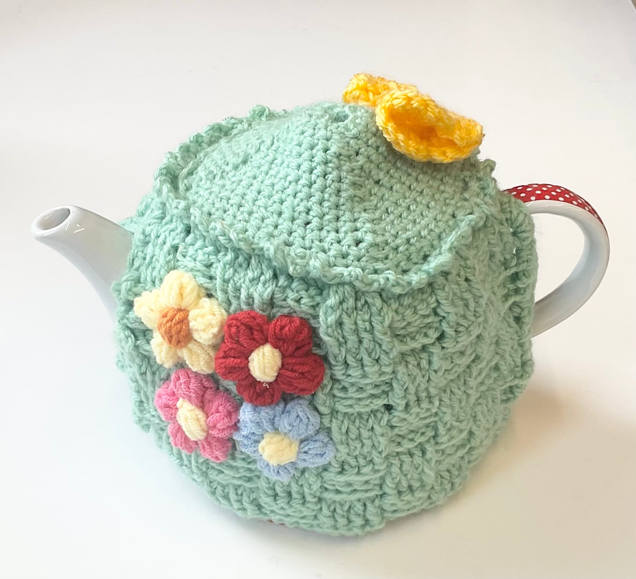 Teapot with a green knitted cover featuring colorful flowers on a white background