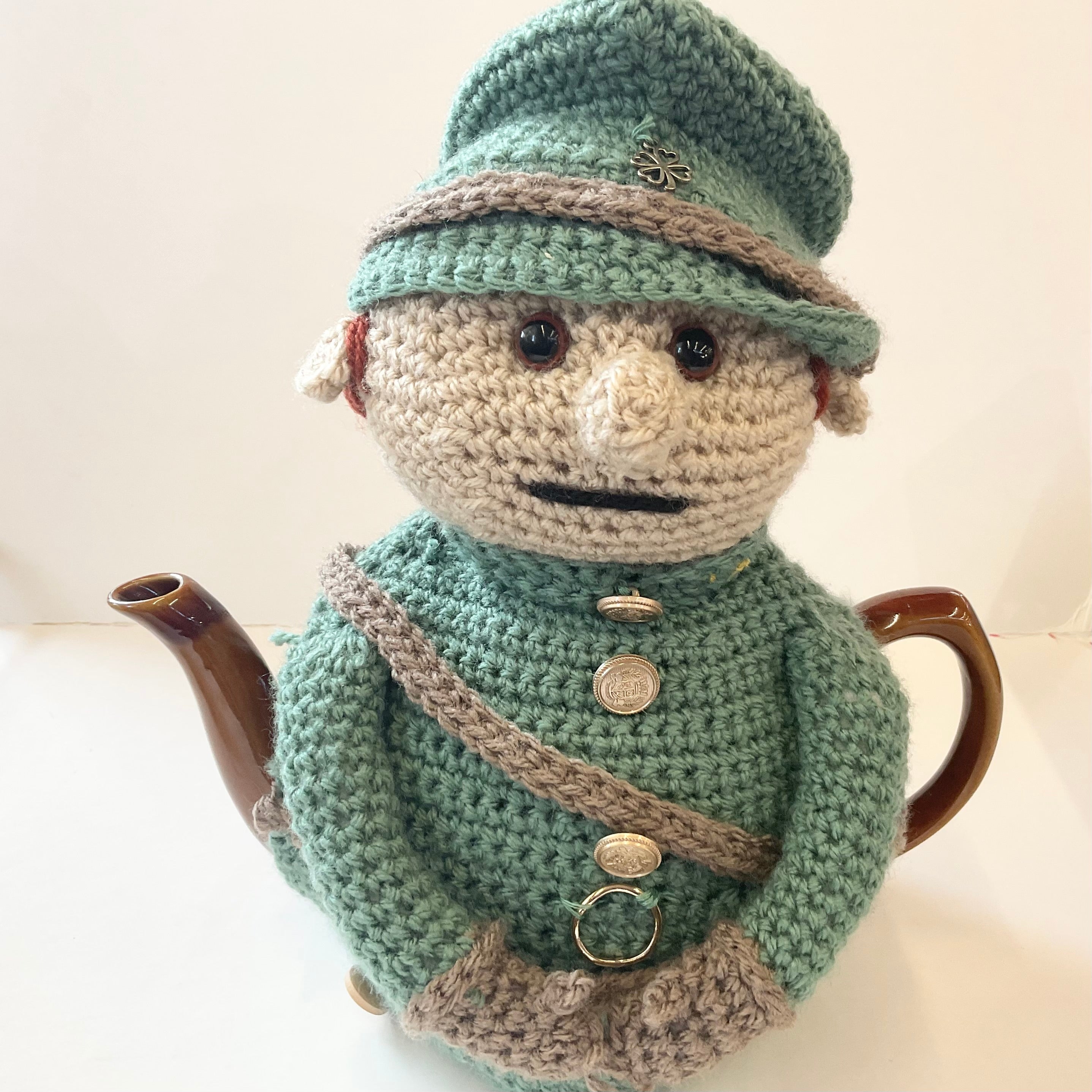 A crocheted doll-shaped teapot featuring a mint green and beige outfit with a hat, face, and brown handle, photographed against a plain light background.