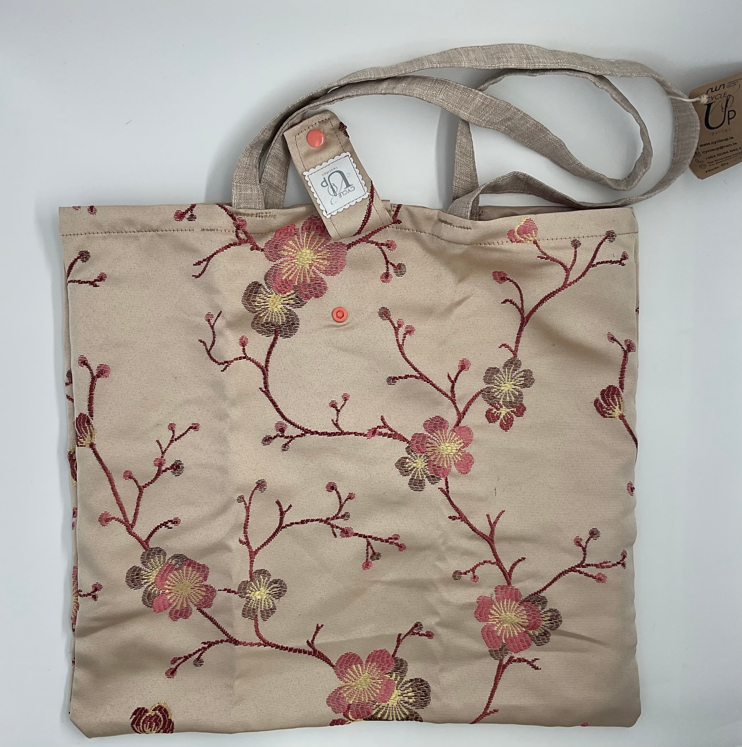 A beige foldable shopping tote bag with a floral pattern in shades of pink and red, featuring brown handles.