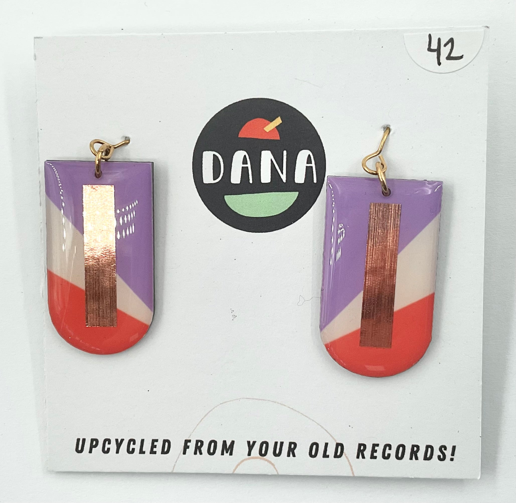 A pair of earrings made from upcycled vinyl records, predominantly red and purple in color, with a gold-colored bar at the top, displayed on a white background with the brand name 'DANA' above.