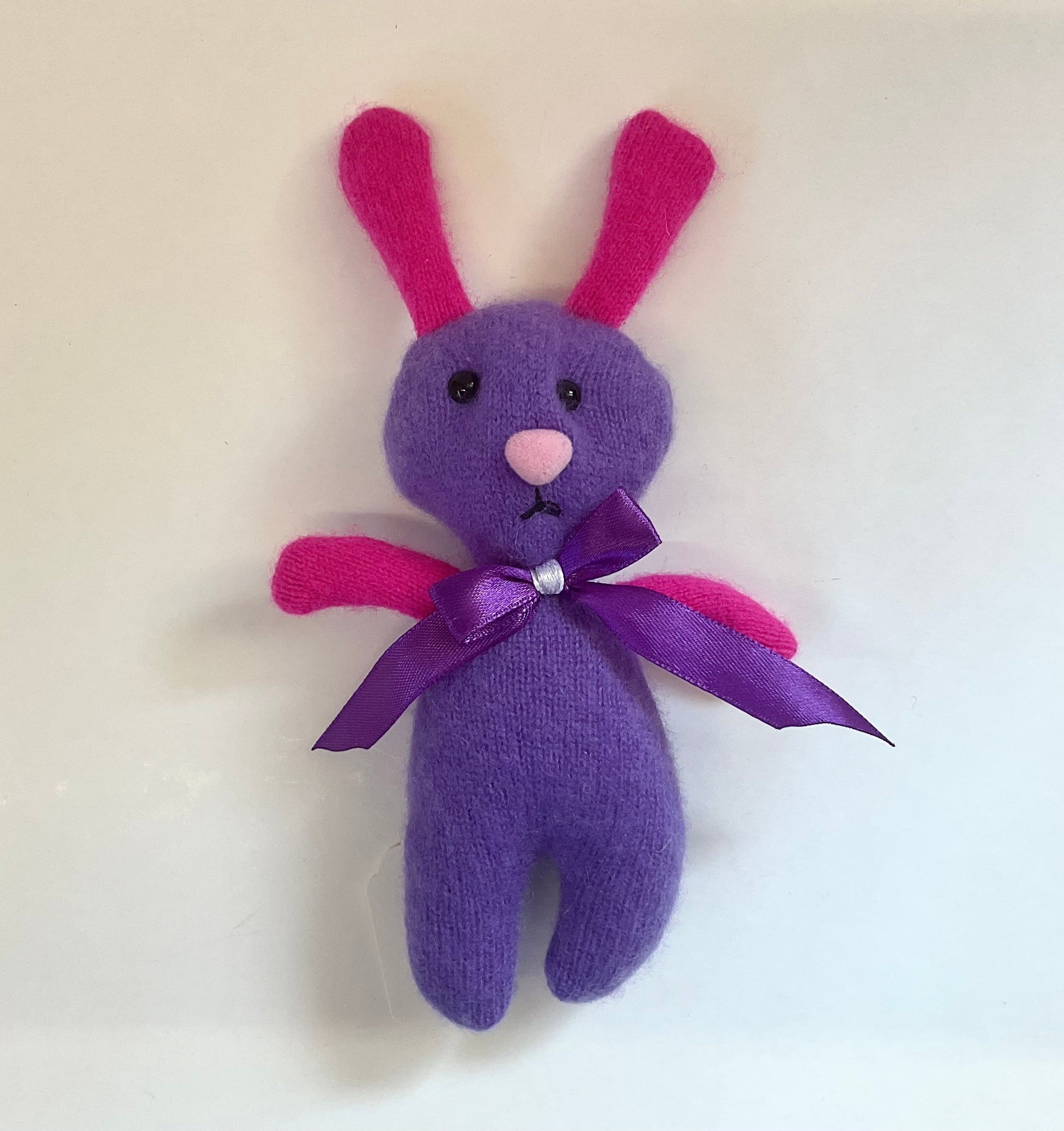 Purple stuffed bunny with pink ears and arms, and a purple bow, on a white background