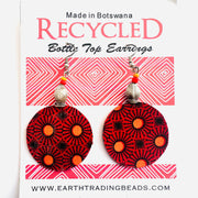 A pair of recycled bottle top earrings with pink, orange, and black geometric patterns, displayed on a card with 'Made in Botswana' text and the brand name 'Earth Trading'.