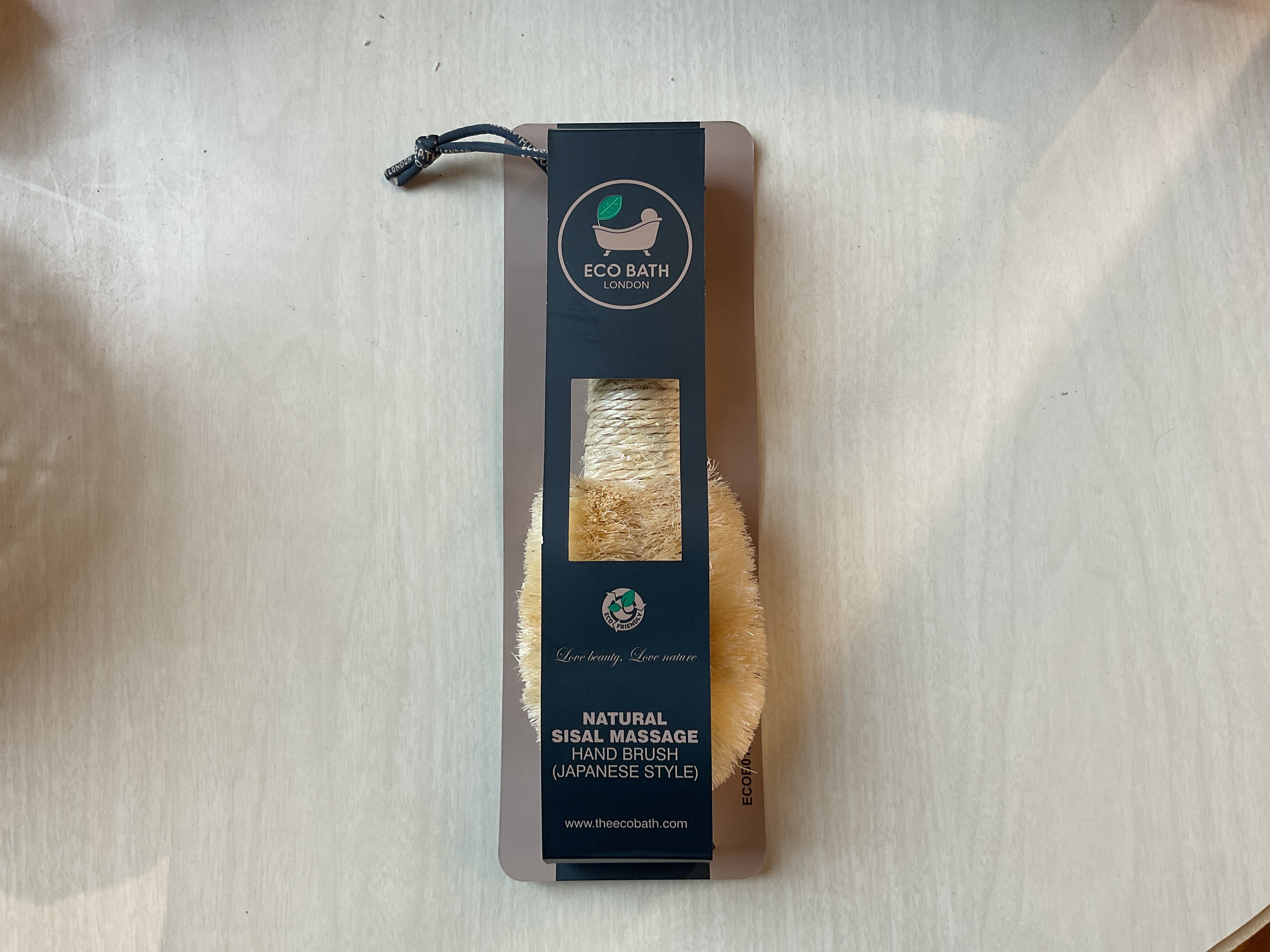 A natural sisal massage hand brush packaged in a brown paper bag with a black label featuring the brand name and product details.