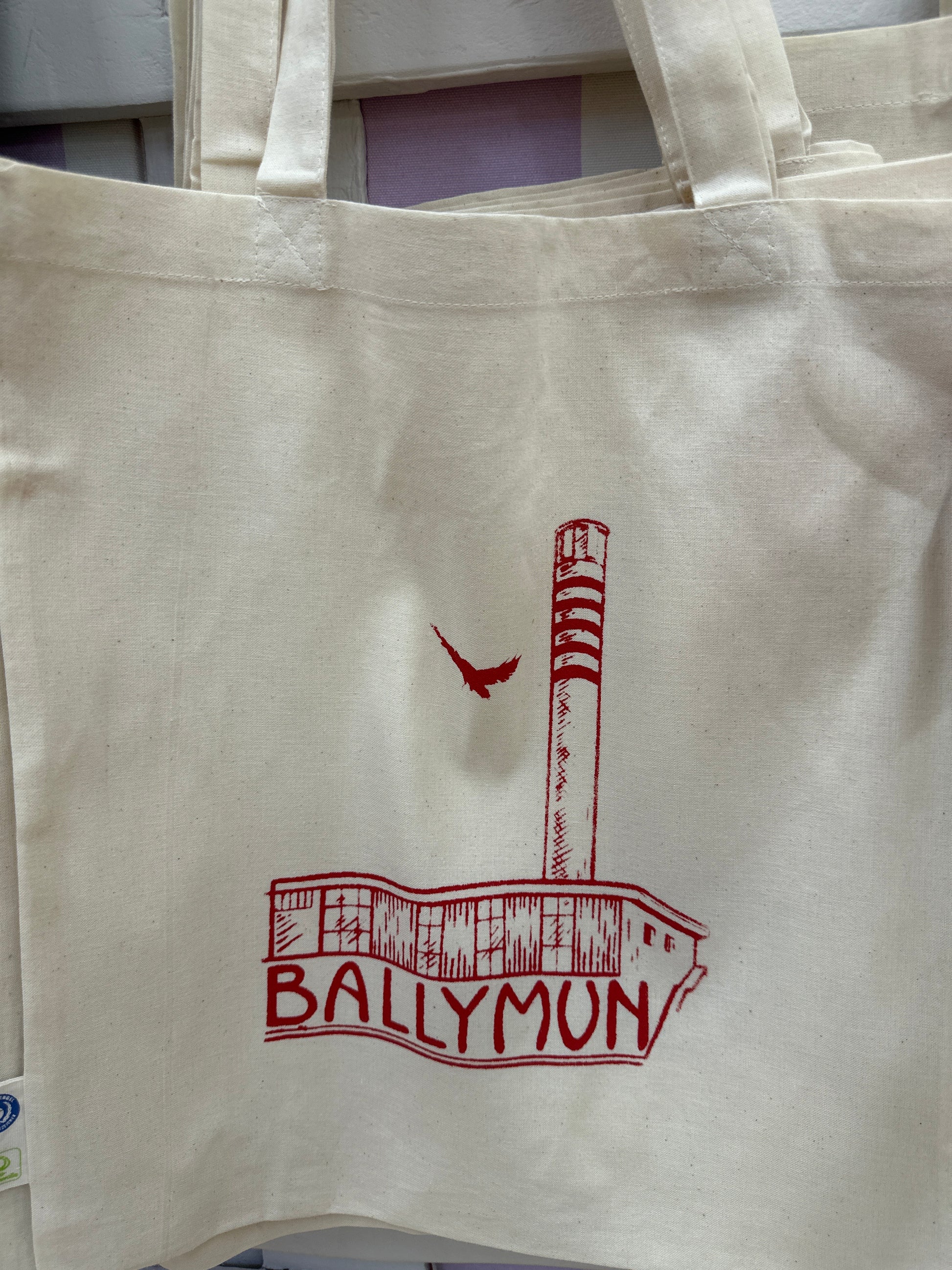 A white cotton tote bag with red text and a graphic that reads 'BALLYMUN' on the front with a print of the Rediscovery Centre and a bird.