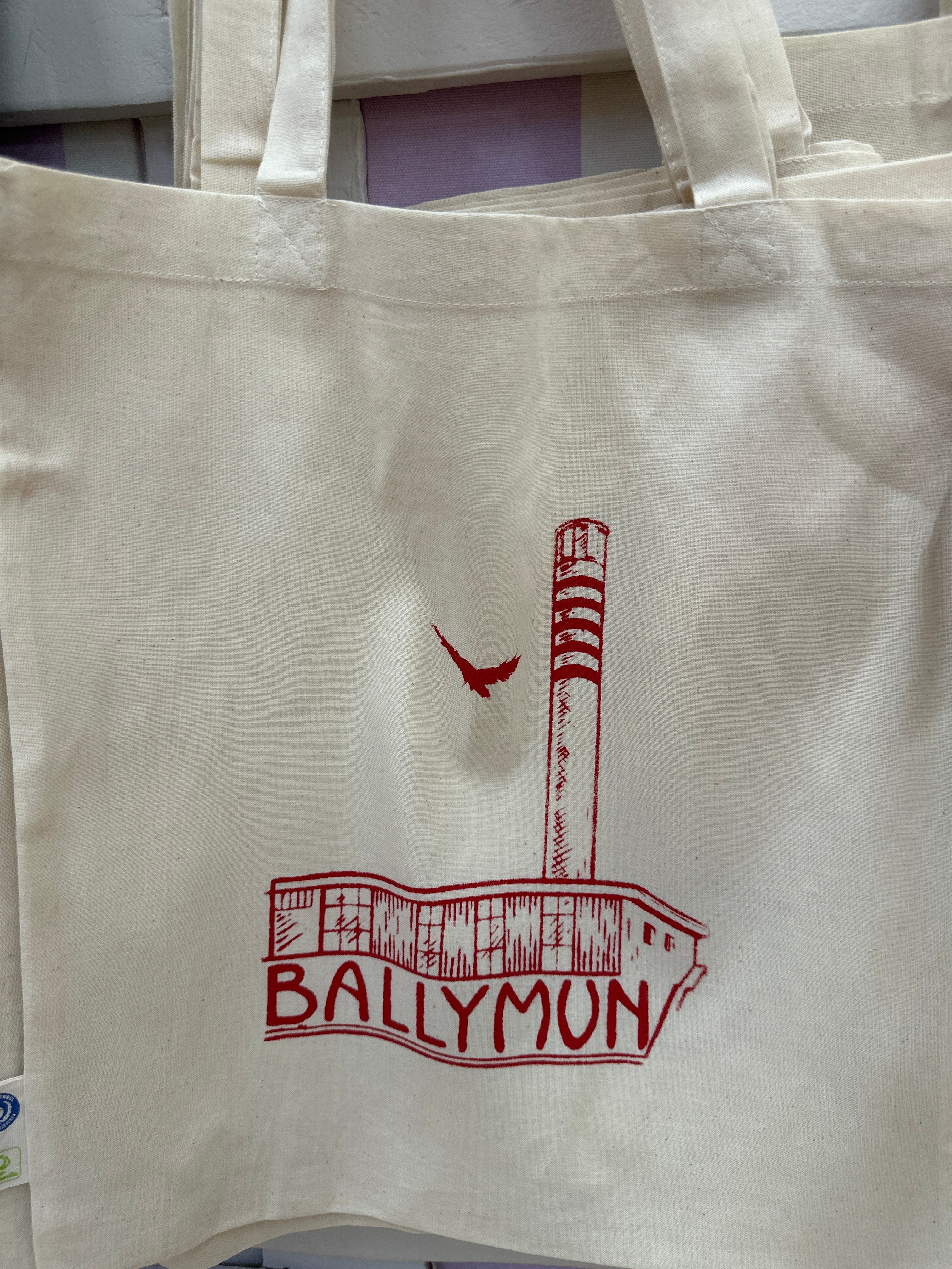 A white cotton tote bag with red text and a graphic that reads 'BALLYMUN' on the front with a print of the Rediscovery Centre and a bird.