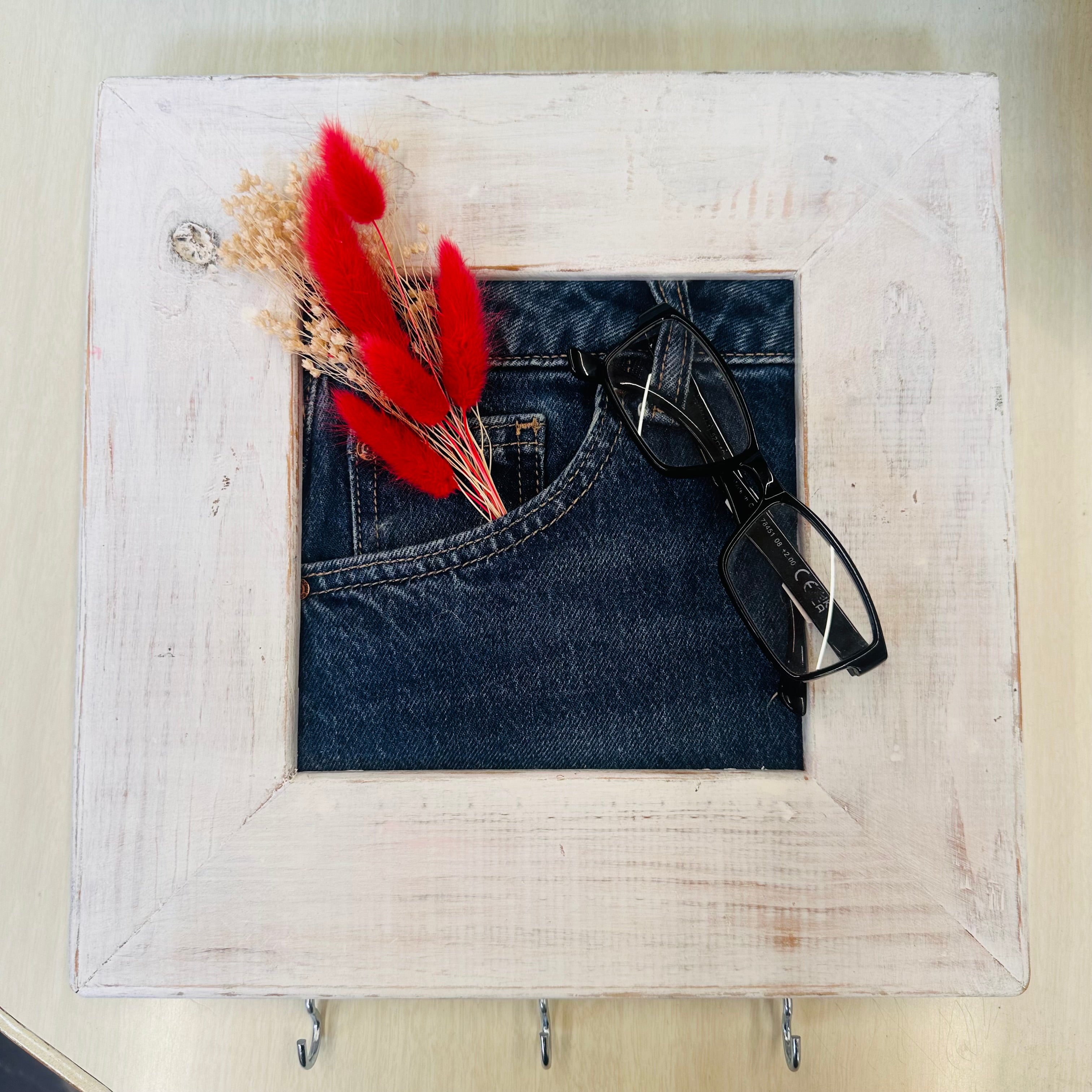 A square white distressed wooden frame with three hanging hooks at the bottom, styled with a folded pair of dark blue jeans, red decorative feathers, and black eyeglasses on a light wood surface.