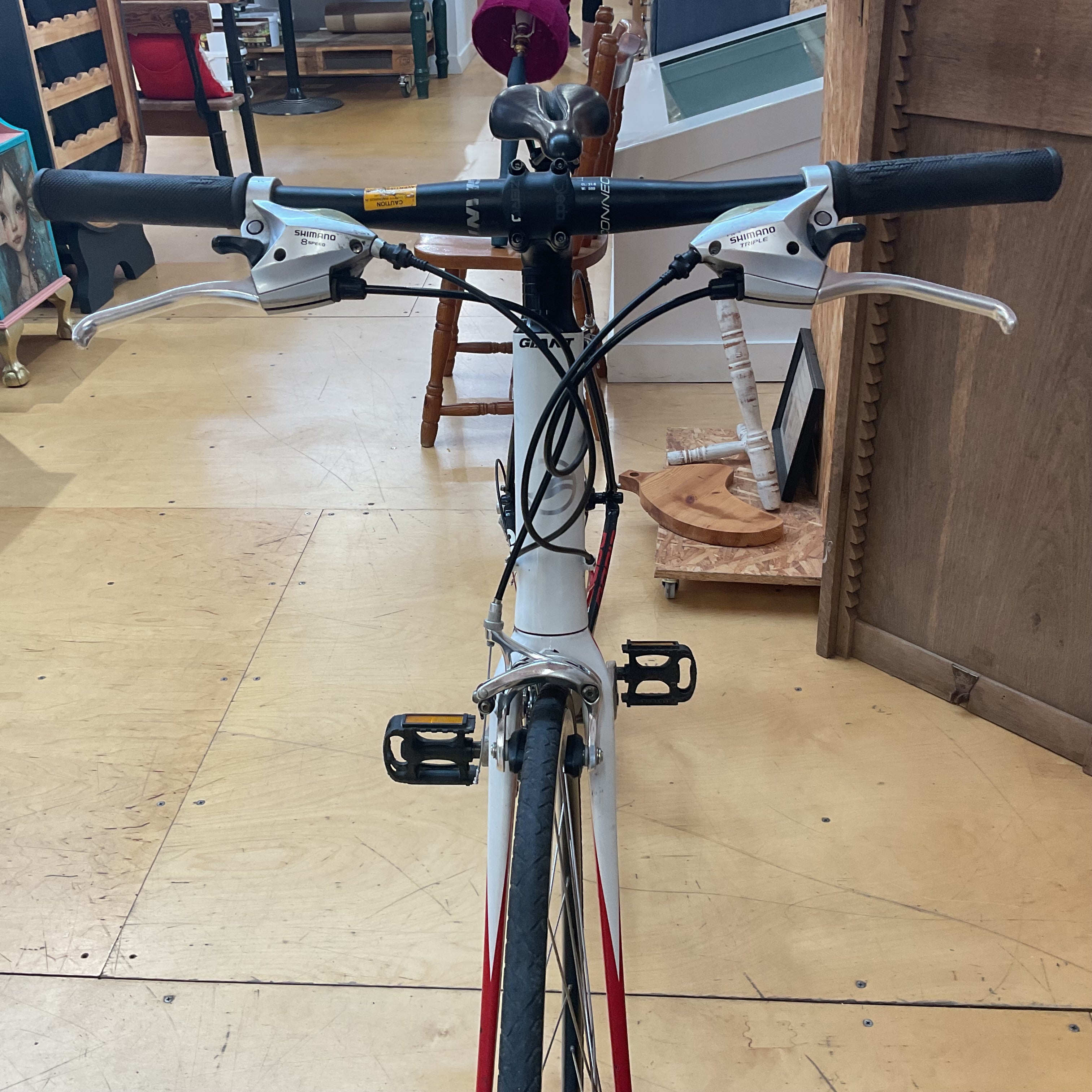 A white bicycle with red and black accents, viewed from the front in a shop setting with wooden floors and furniture in the background.