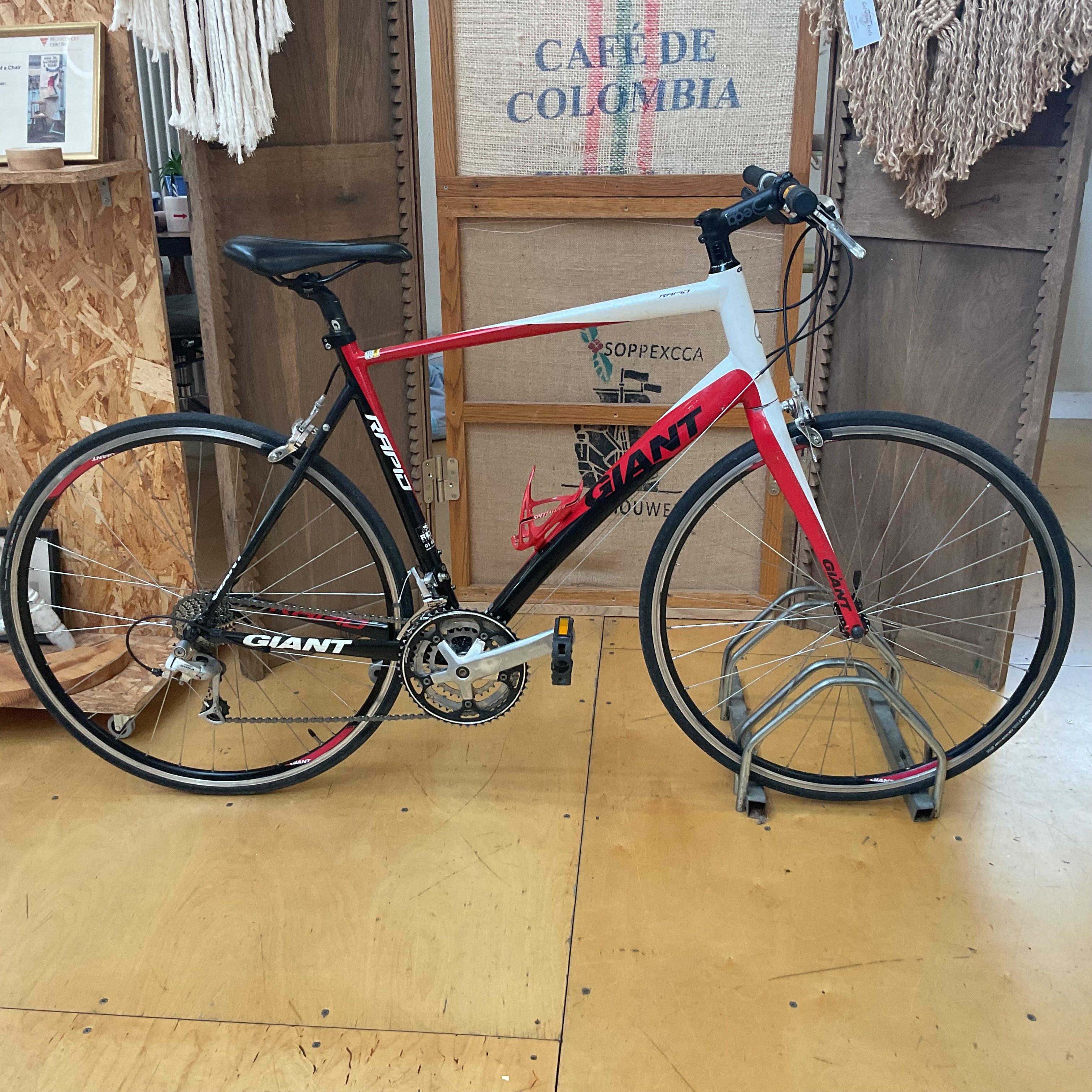 A Giant road bicycle with a red, white, and black frame, displayed on a metal stand in a shop setting with wooden floors and burlap backings.