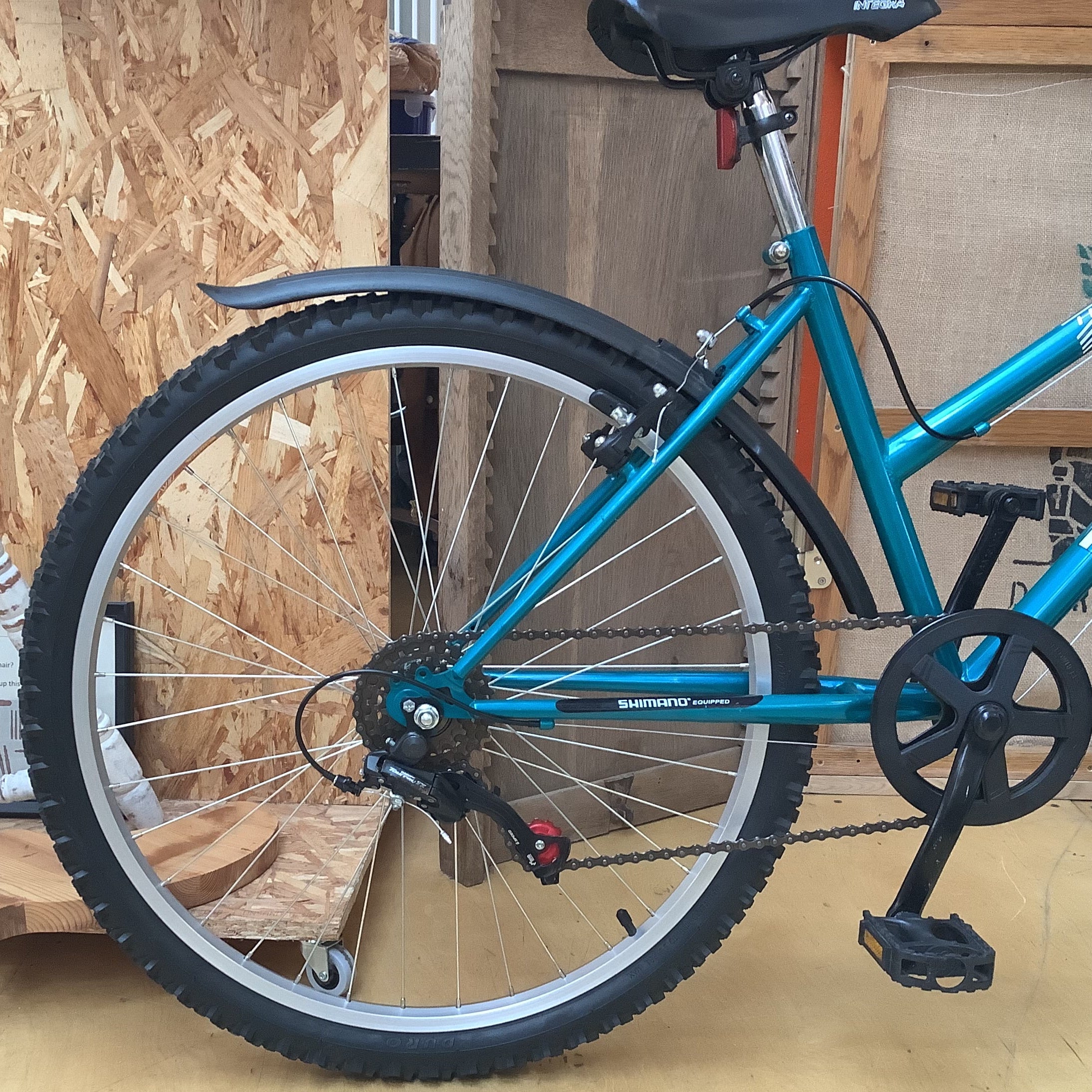 A teal Shimano bicycle with black tires, fenders, saddle, and pedals, photographed indoors against a background of plywood panels and wood boards.
