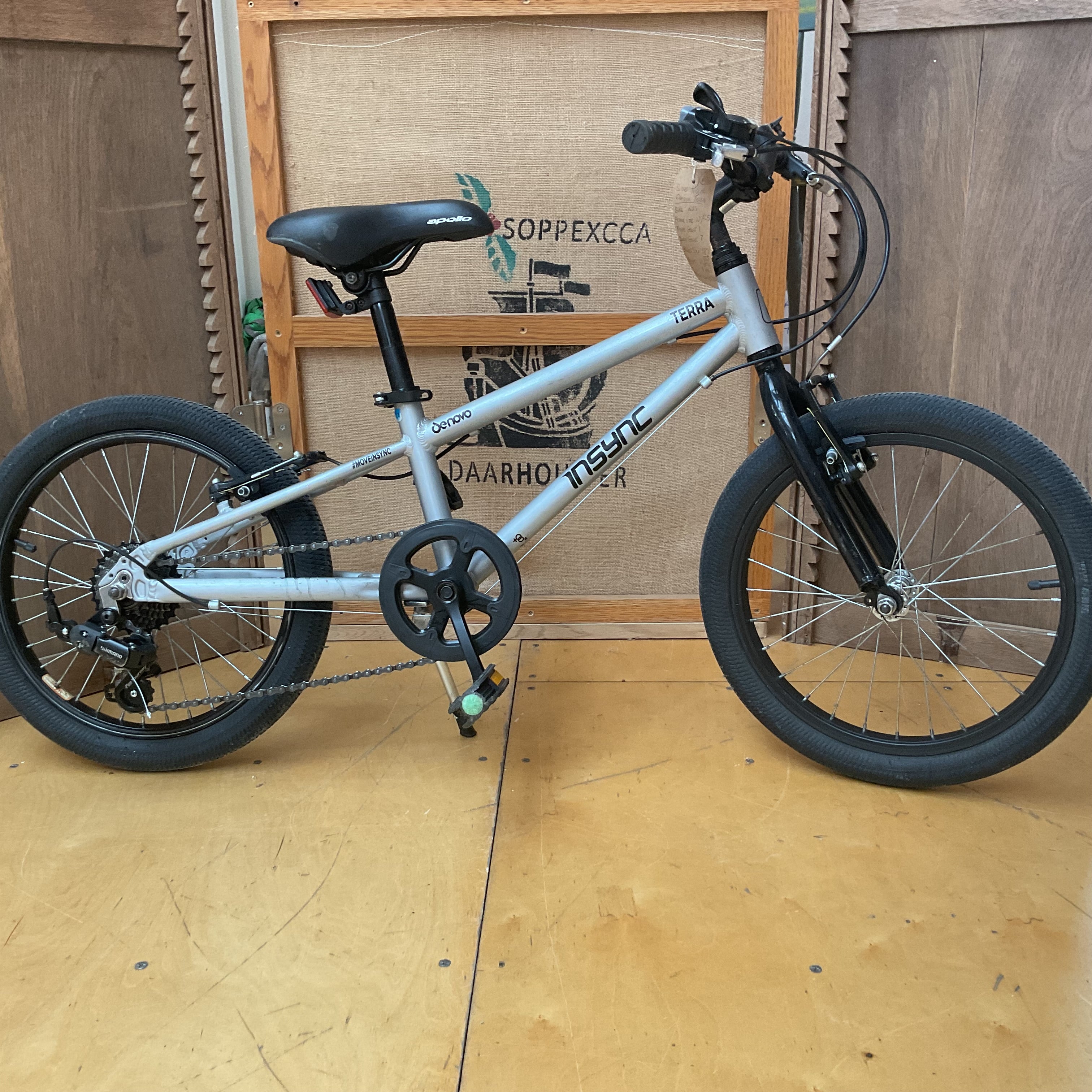 A light silver BMX bicycle with black tires, a black saddle, and a black front fork, displayed on a wooden floor in front of a wooden crate.
