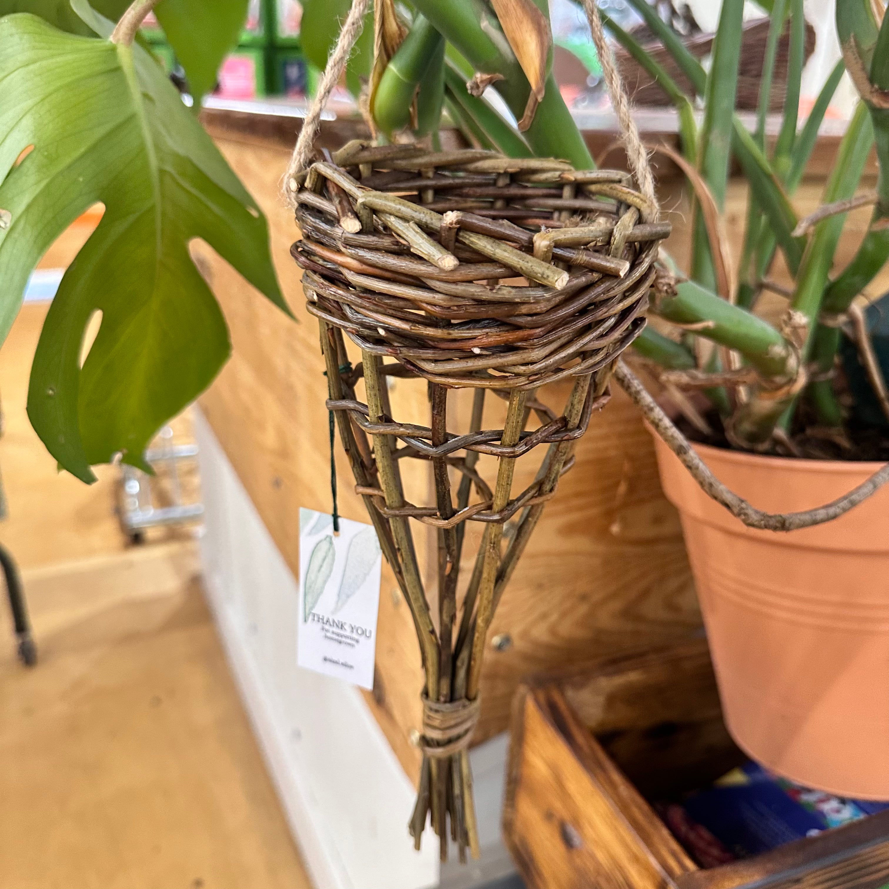 A woven willow bird feeder with visible twine and hanging leaves, displayed in a garden setting.