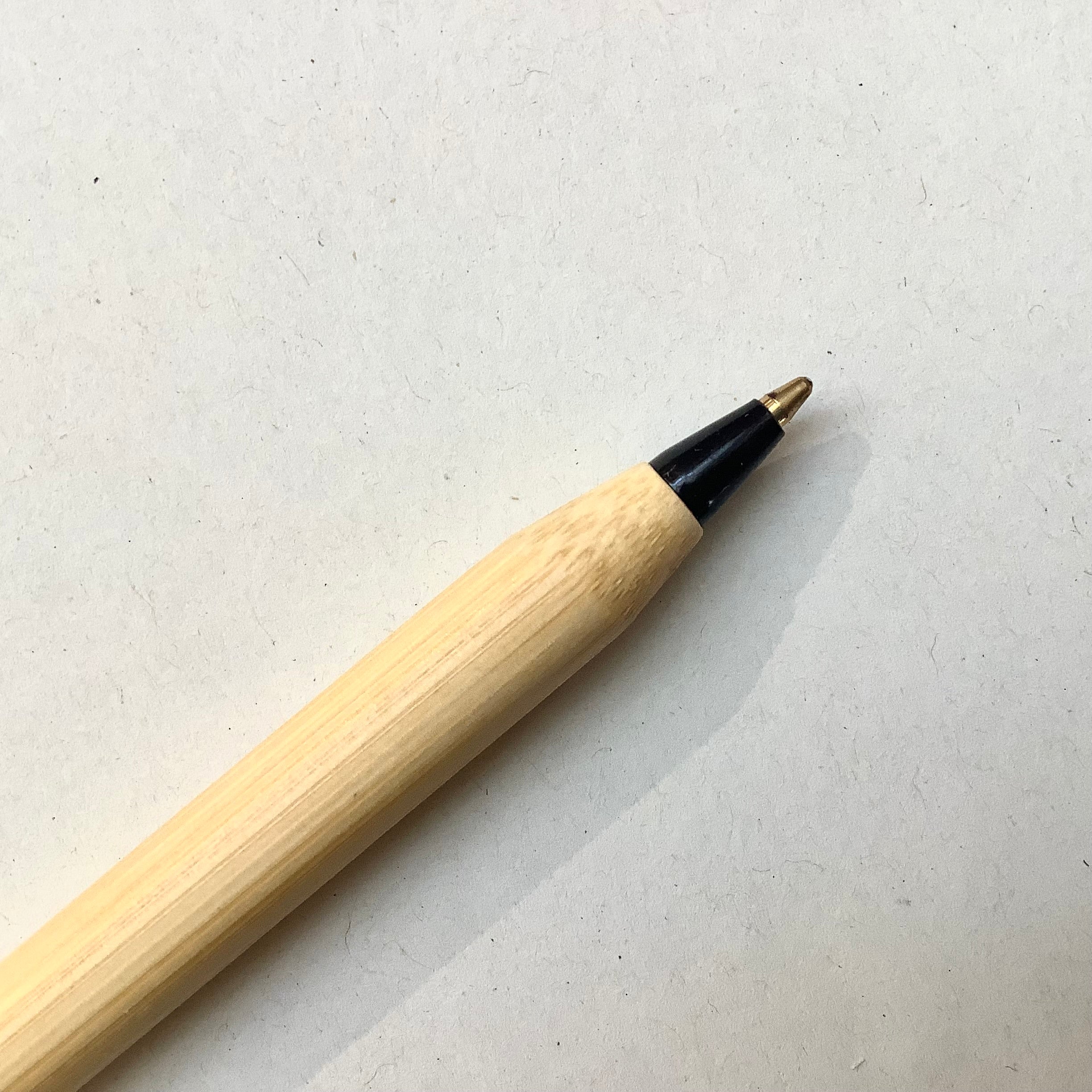 A close-up of a wooden pen with a light natural wood body, a black ferrule, and a gold metal tip, resting on a textured off-white surface.