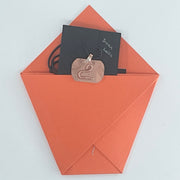 A copper-toned metal pendant necklace with a stylized swan cutout, shown tucked inside an orange envelope with a black card labeled 'Swan Lake' on a light gray background.