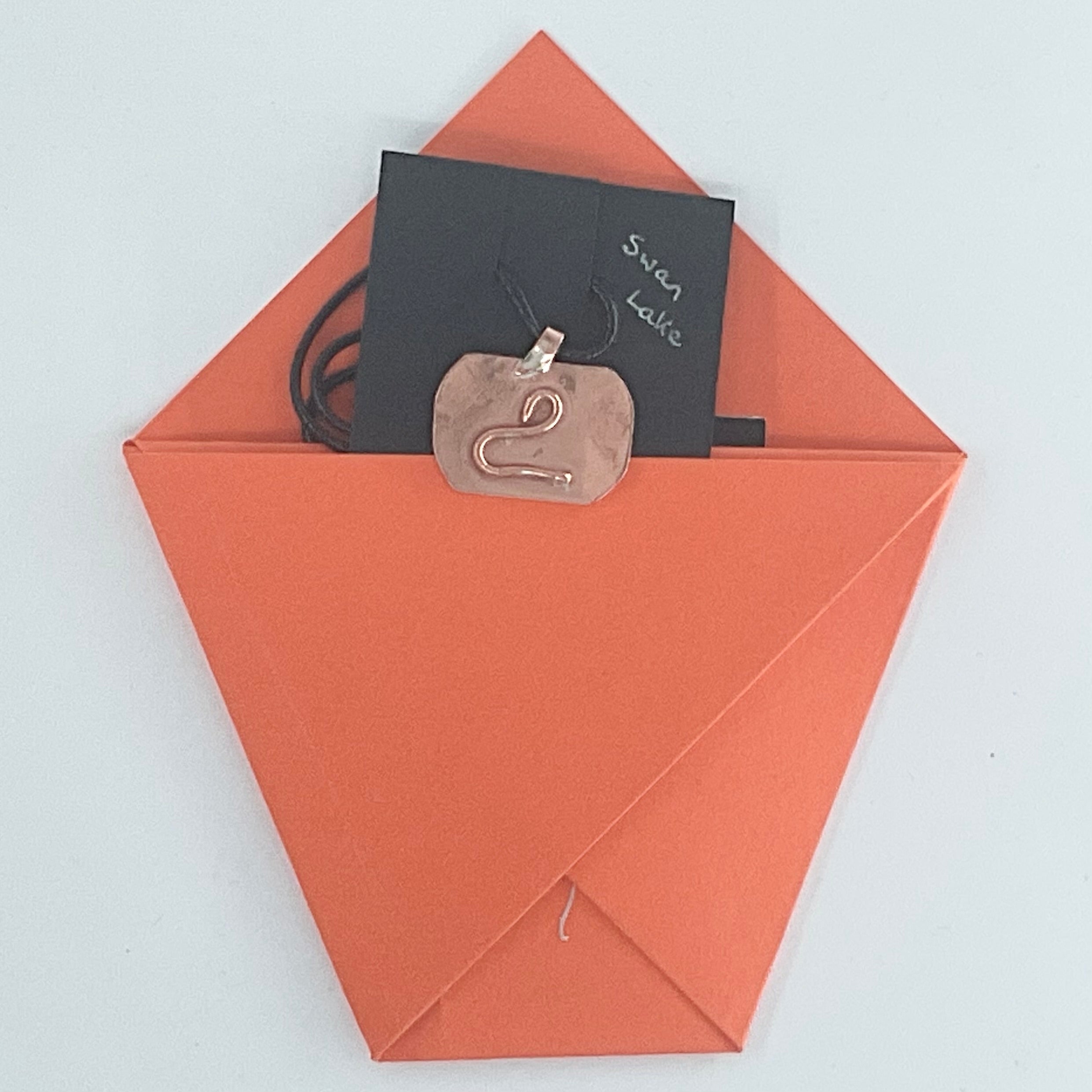 A copper-toned metal pendant necklace with a stylized swan cutout, shown tucked inside an orange envelope with a black card labeled 'Swan Lake' on a light gray background.
