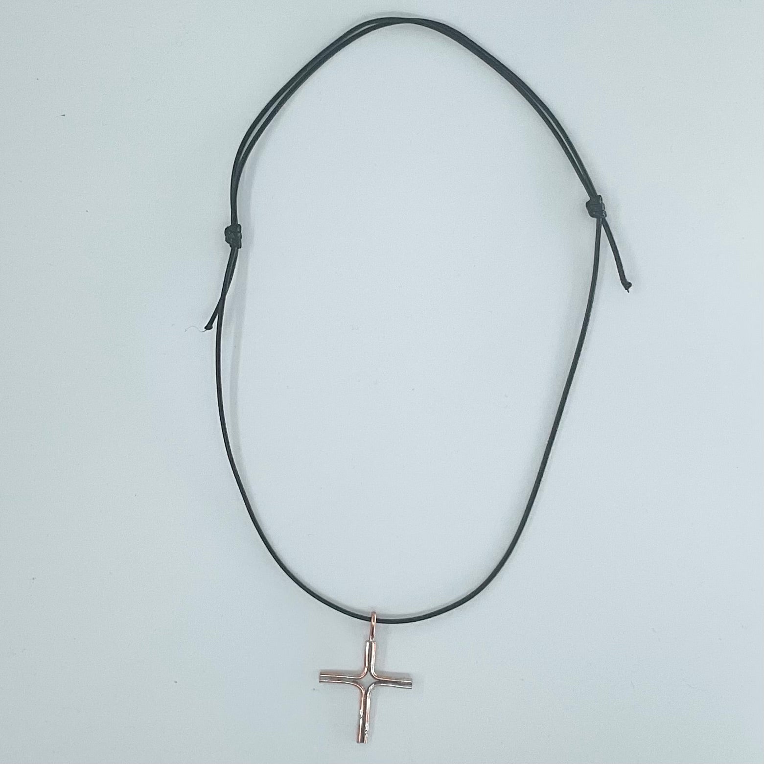 A black adjustable cord necklace with a small rose gold cross pendant, displayed on a light gray background.