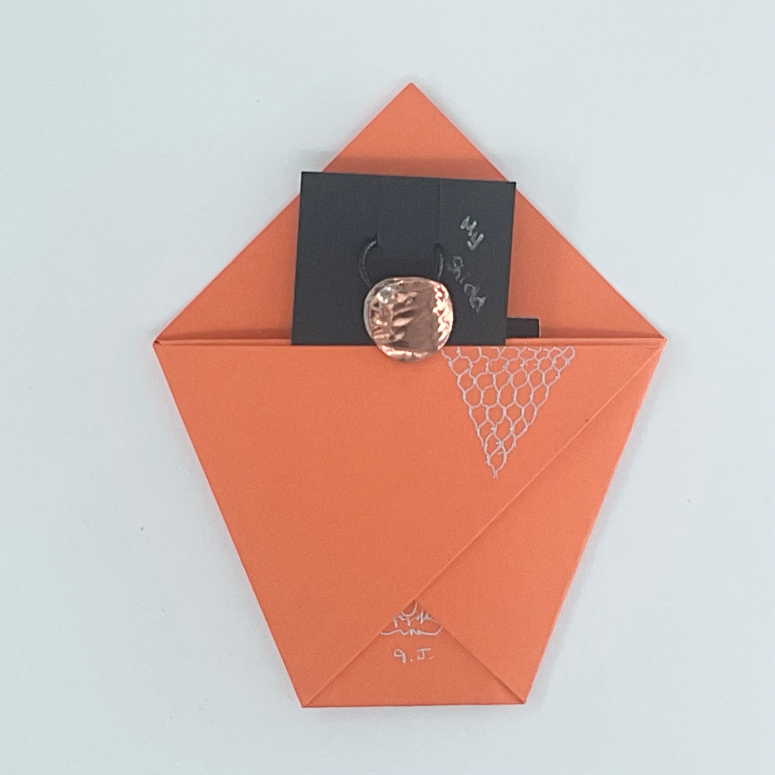 An orange paper envelope or pocket-style card holder with a black card partially pulled out and a round metallic rose-gold coin resting on top, photographed on a plain light background.
