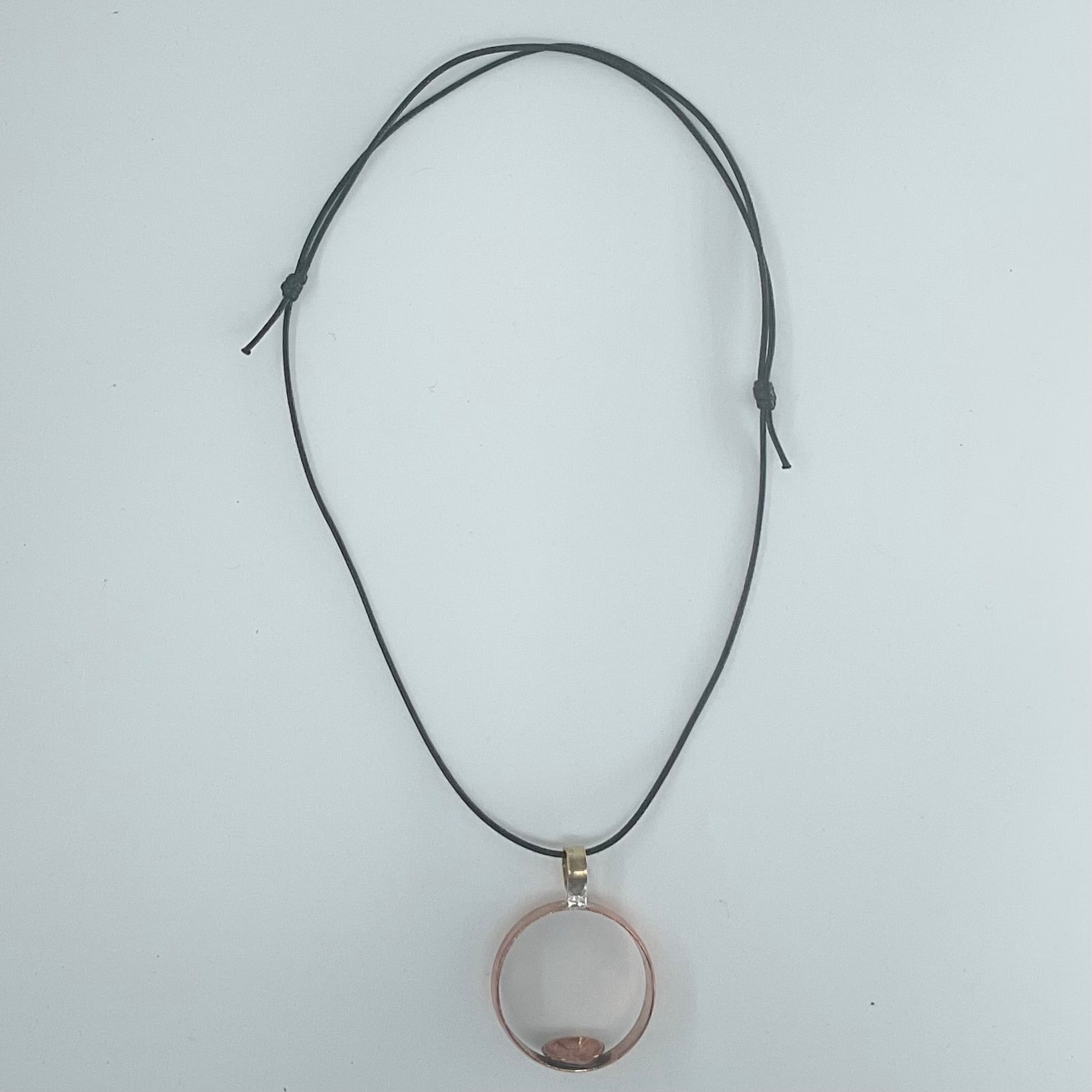 A black adjustable cord necklace with a circular pendant containing a small pinkish-brown shell, displayed on a light gray background.