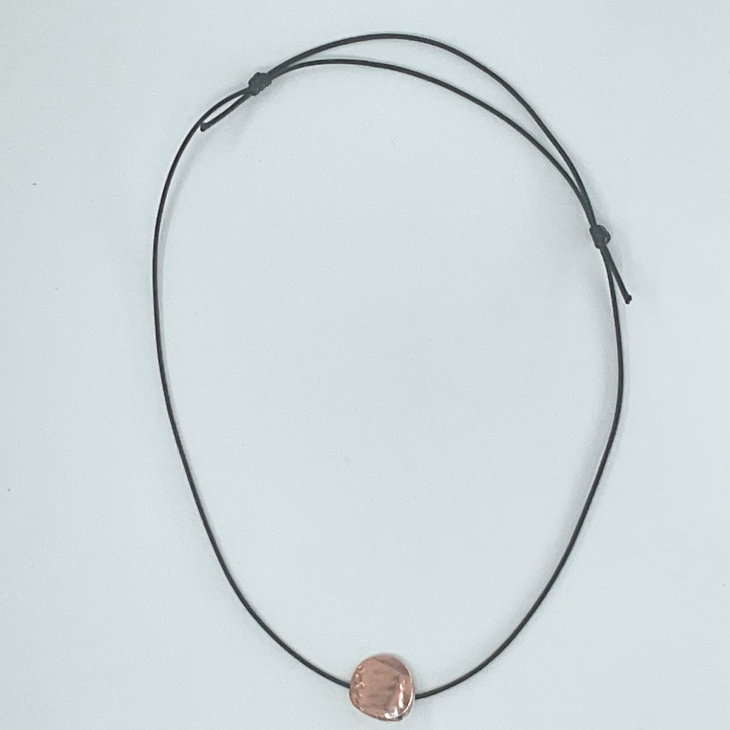 A minimalist black cord necklace with a small, textured rose gold disc pendant, displayed on a clean white background.