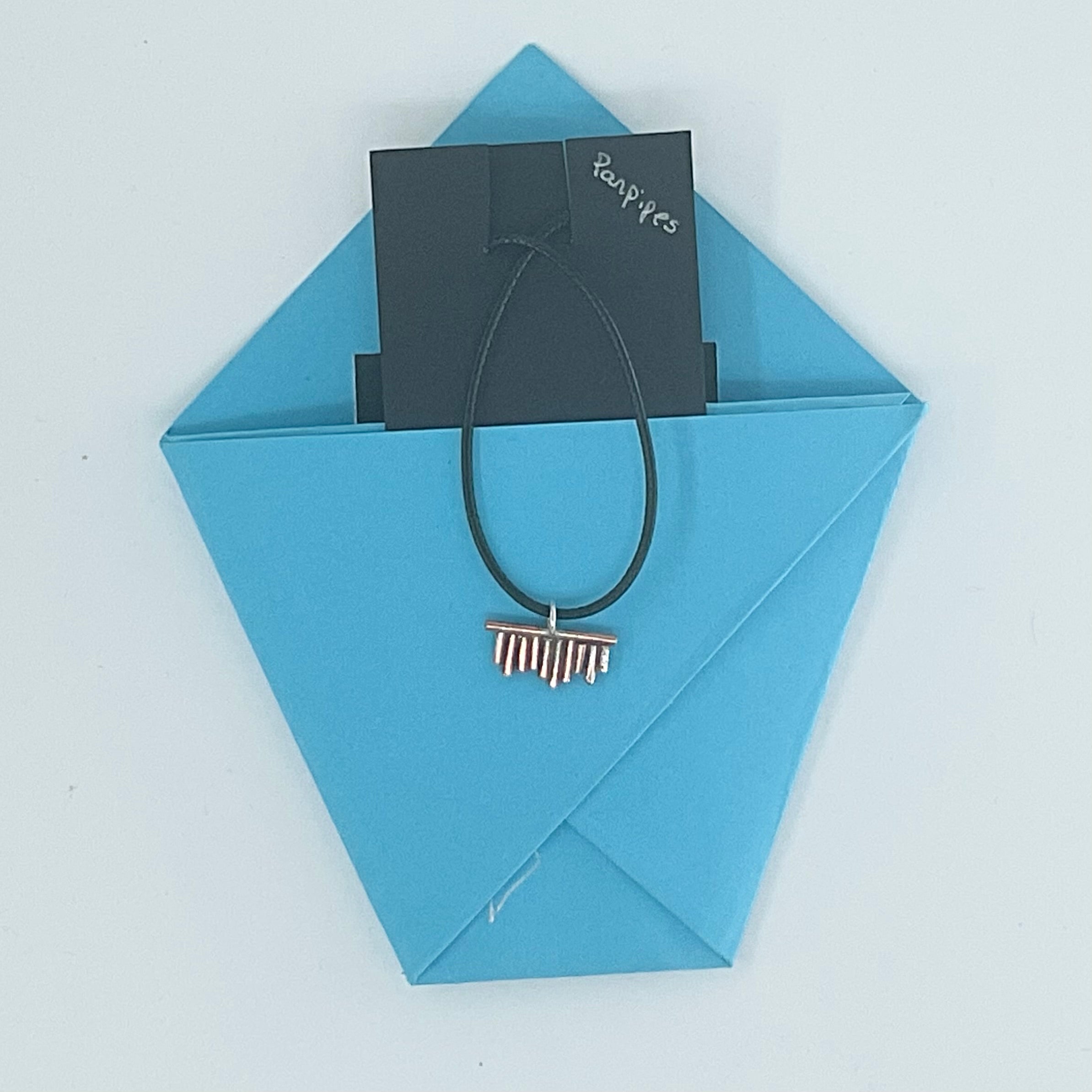 A black cord hair tie with a small metallic barrette-style end, presented on a light blue envelope with a black card labeled 'Bangles' tucked behind it, on a white background.