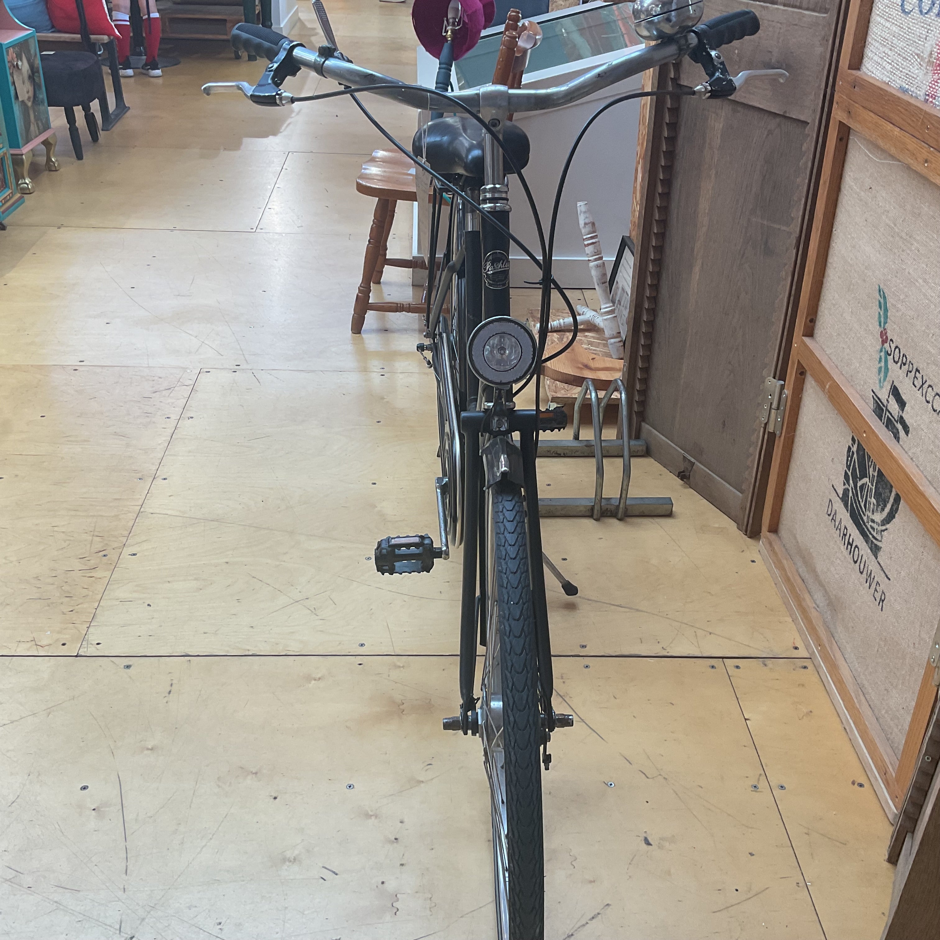 A black vintage-style bicycle with a round front headlight and silver handlebars, photographed indoors on a light wood floor. A person's legs are partially visible in the background near a wooden stool and door.