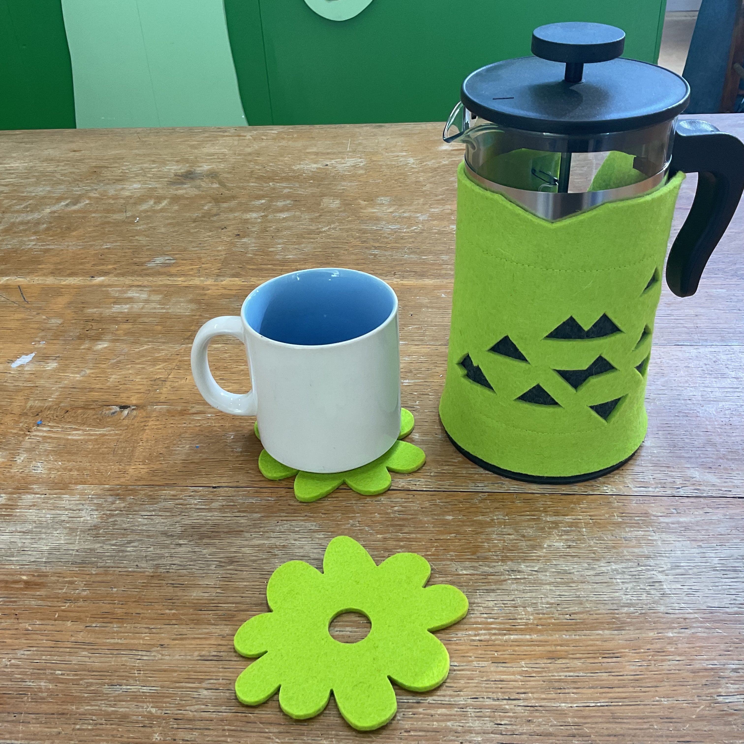A bright green felt flower-shaped coaster and matching coaster under a white ceramic mug with a blue interior, alongside a glass French press with a green felt sleeve on a wooden table.