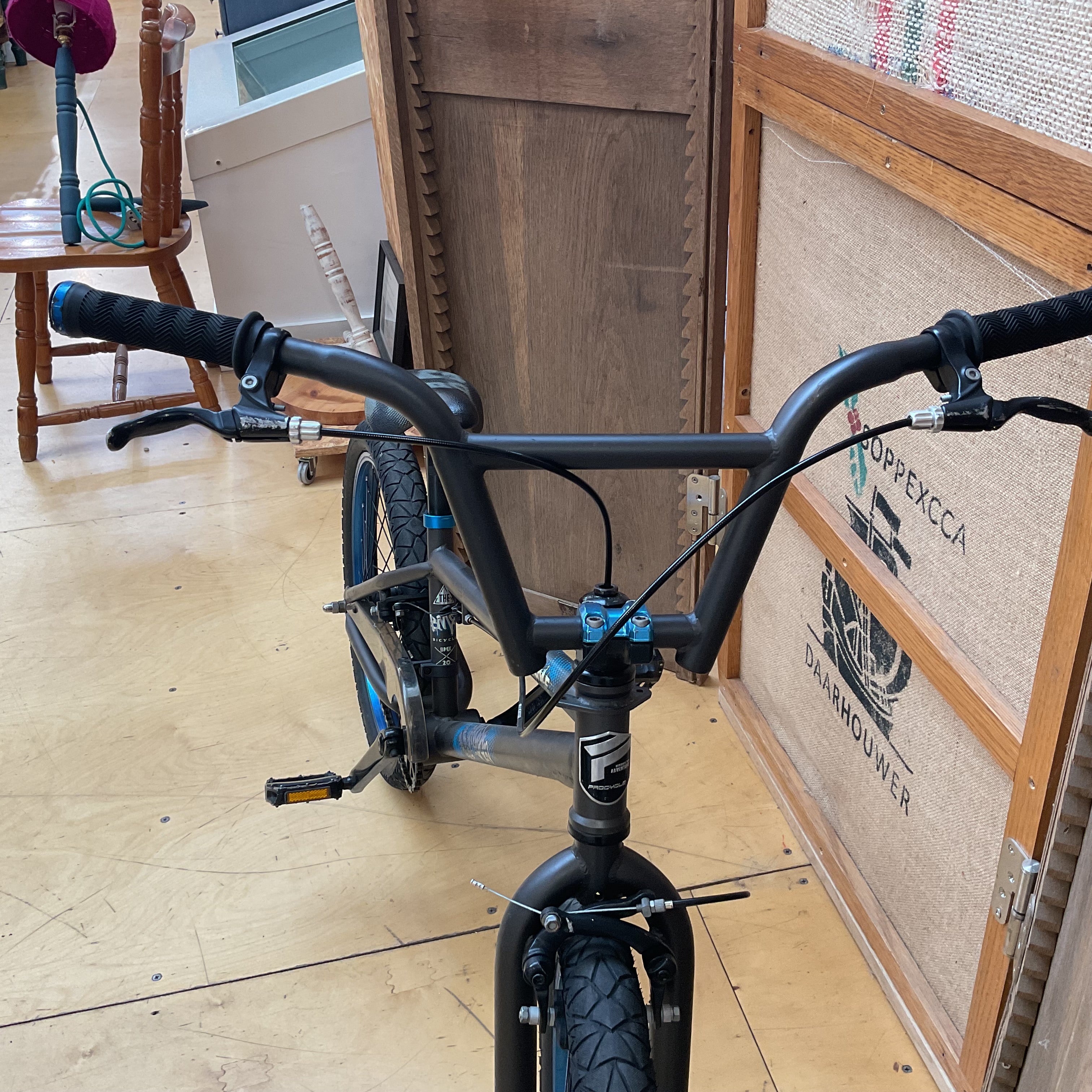 A close-up view of a BMX bike's handlebars and front fork, featuring a matte black frame with bright blue accents on the headset and seatpost. The bike is photographed indoors on a light wood floor next to a wooden crate.