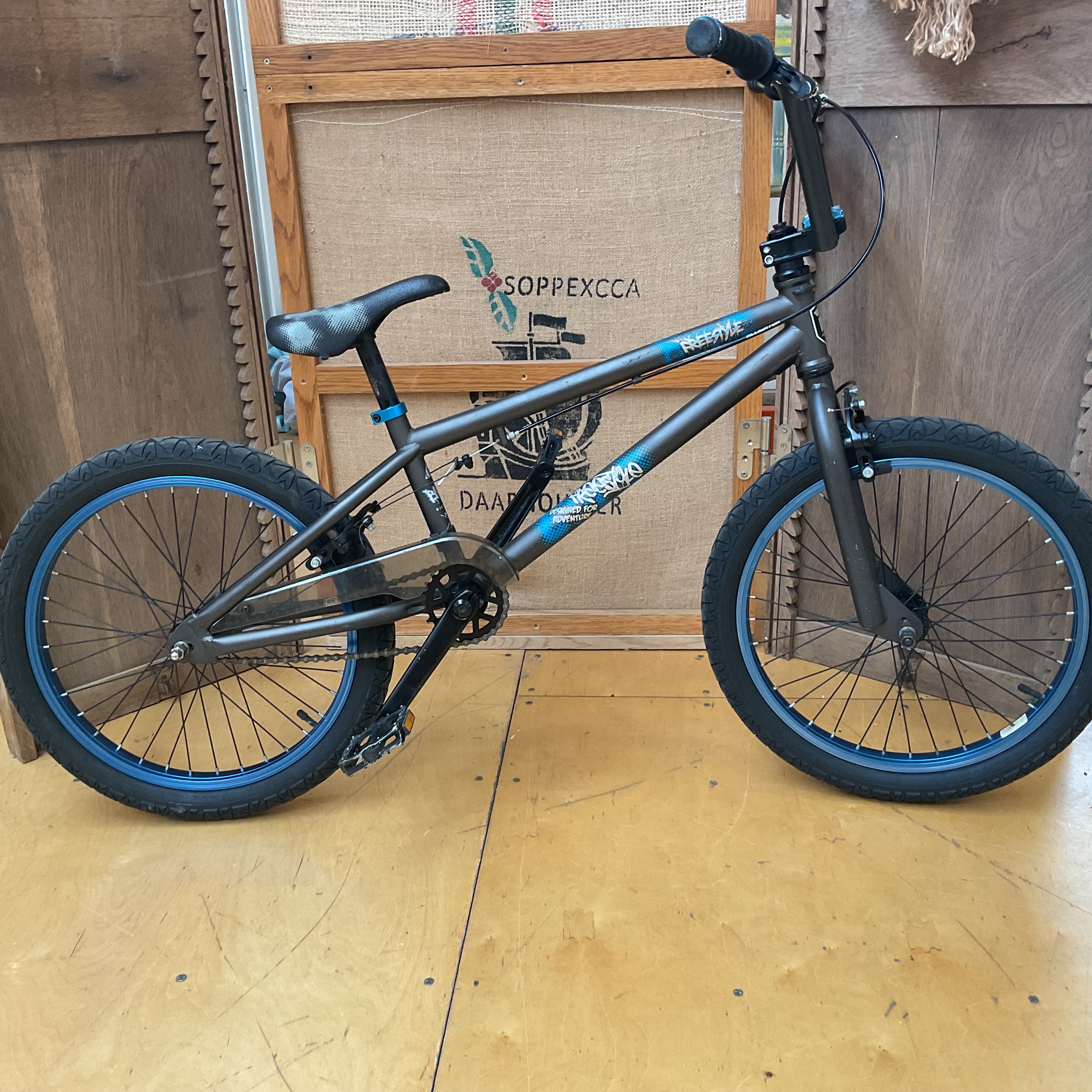 A dark gray BMX bike with blue accents on the rims and frame details, displayed on a wooden floor in front of a wooden wall and a burlap sign.