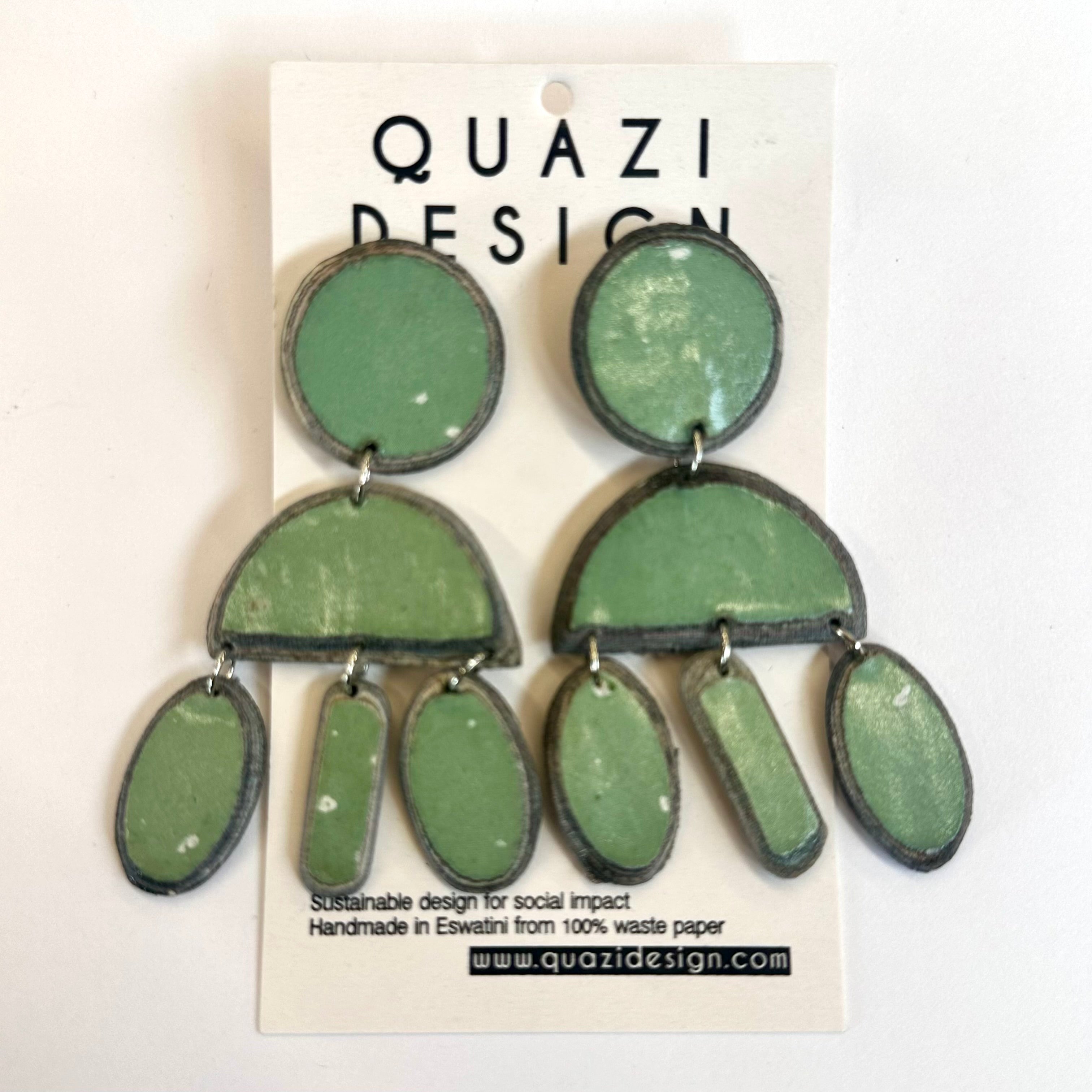 A pair of drop earrings made of recycled magazine paper in green and pink colors, displayed on a card with the brand name 'QUAZI DESIGN'.