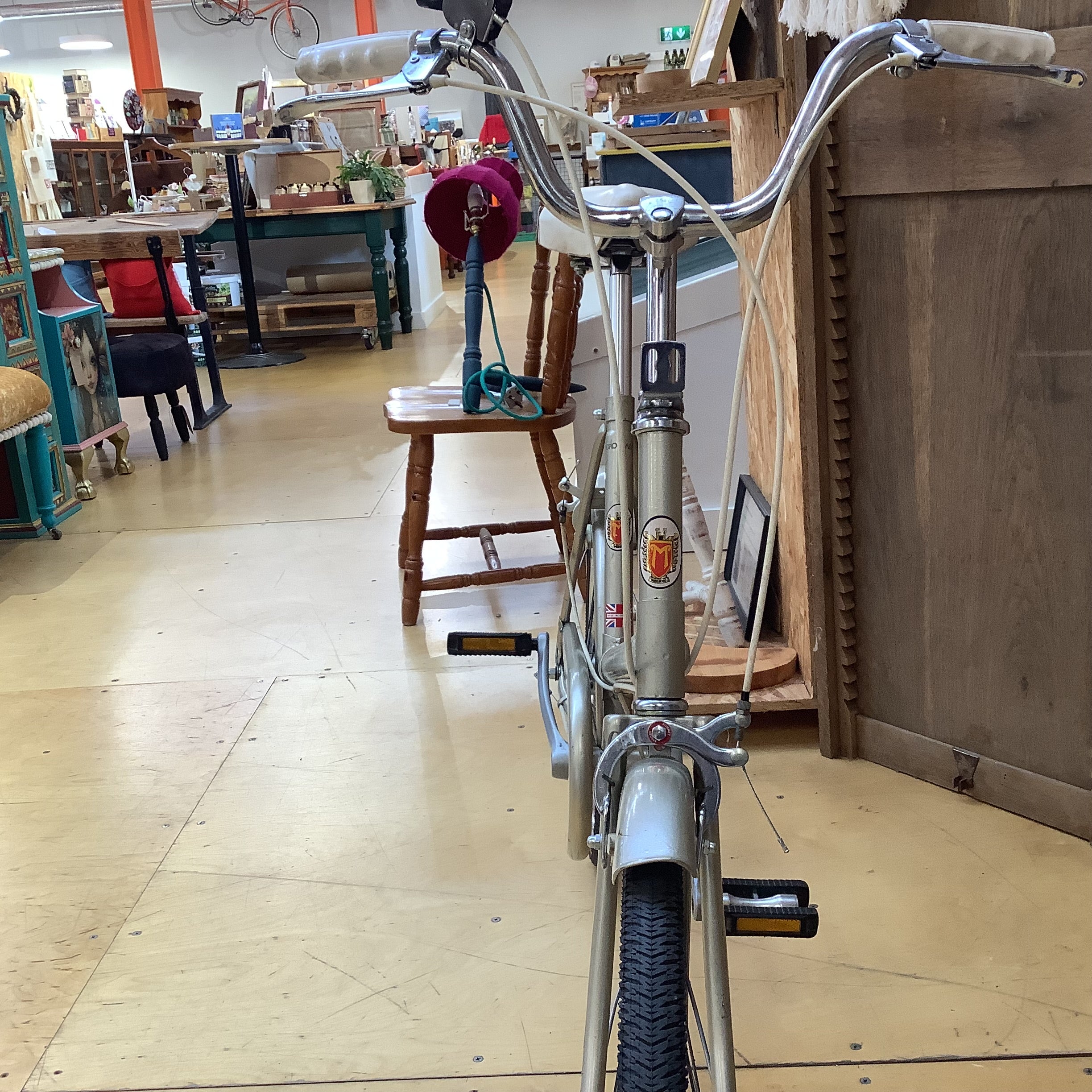 A vintage silver bicycle with high handlebars and a step-through frame, photographed inside a cluttered antique shop or furniture store.
