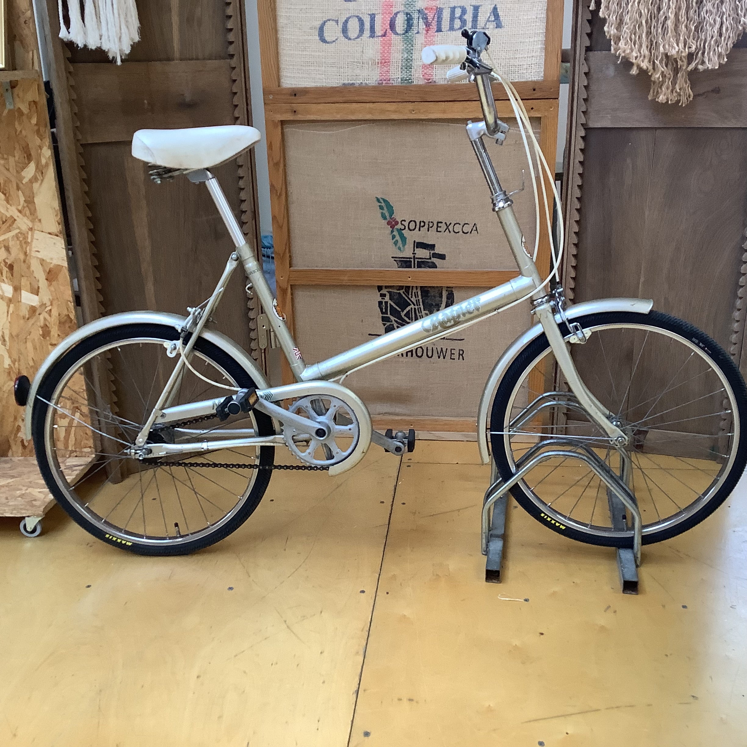 A vintage step-through bicycle with a cream-colored frame, white saddle and fenders, and black tires, displayed on a stand in front of a wooden backdrop with a burlap sign.