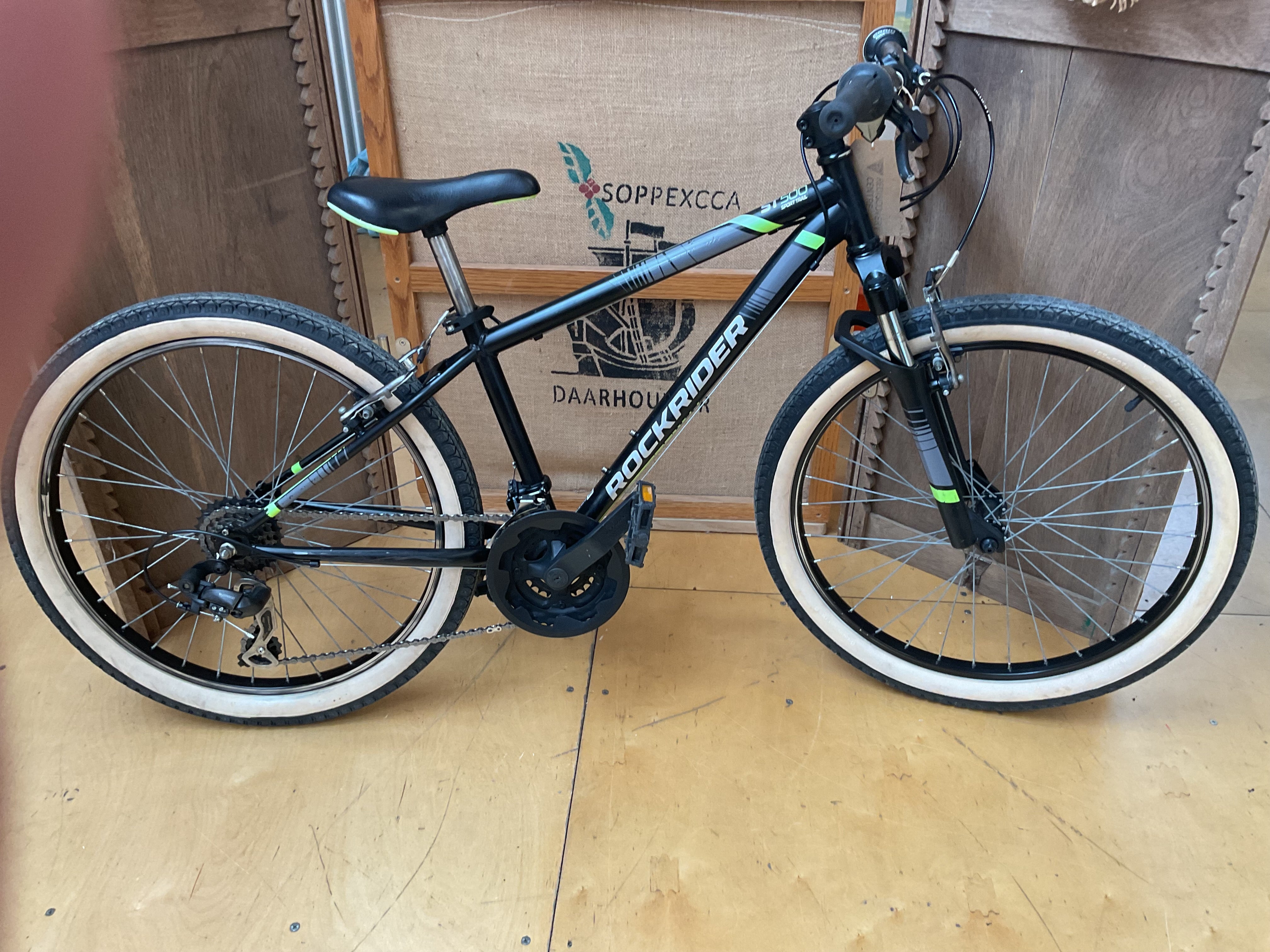 A black Rockrider kids' mountain bike with white-wall tires and green accents, photographed indoors against a cardboard box and wooden wall.