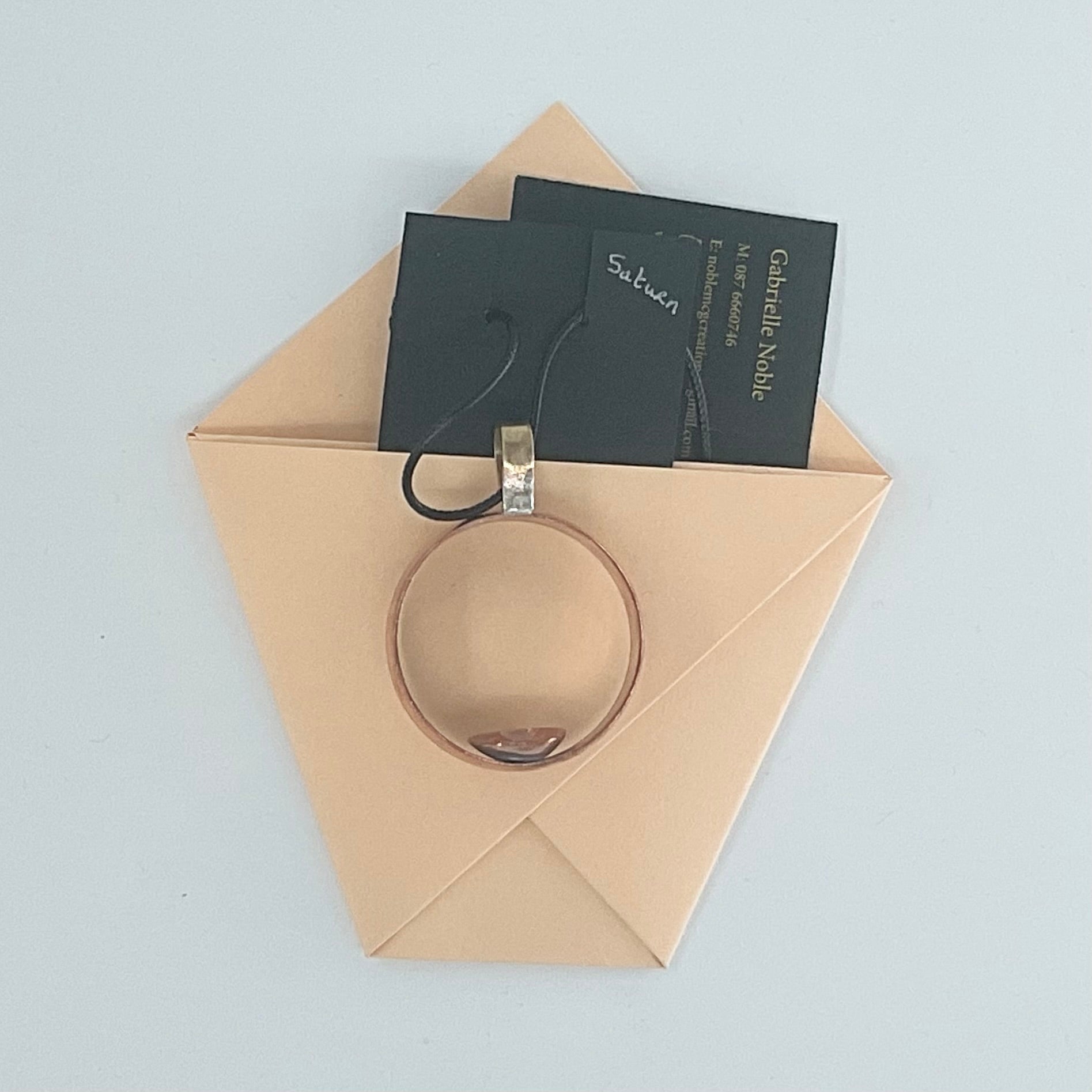 A rose gold ring with a silver-toned bail and black hangtag, displayed inside a beige paper envelope on a light gray background. The tag reads 'Sublime' and 'Catherine Nail'.
