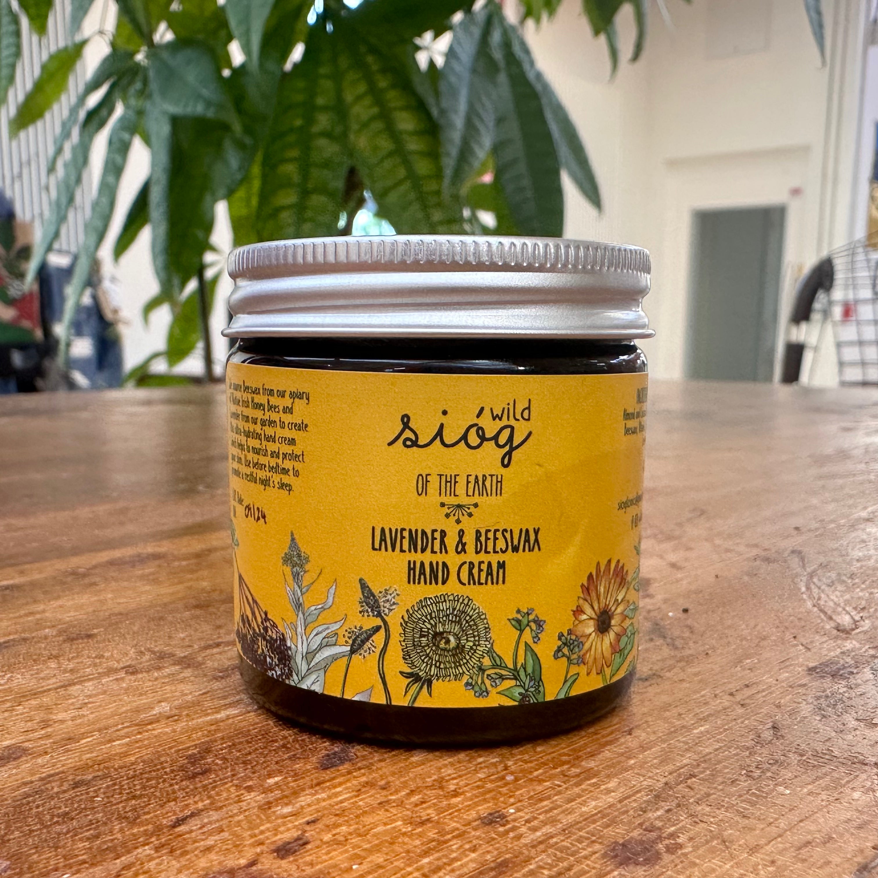 A jar of Sióg of the Earth Lavender & Beeswax Handcream placed on a wooden table with a yellow label featuring floral illustrations.