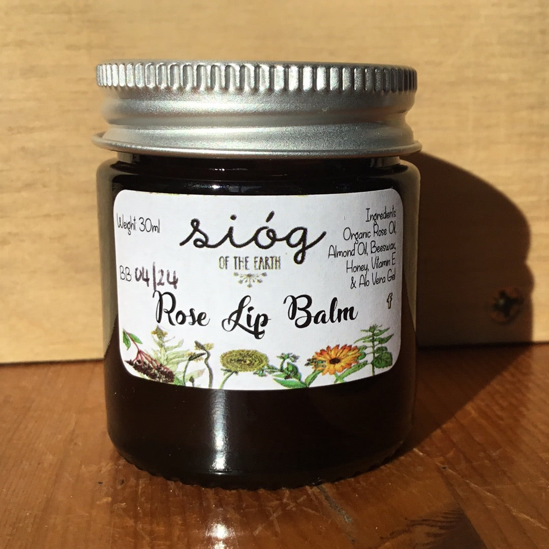 A jar of Sióg of the Earth Rose Lip Balm with a label showing the brand name, product name, and ingredients such as rose oil, almond oil, beeswax, honey, vitamin E, and aloe vera gel.