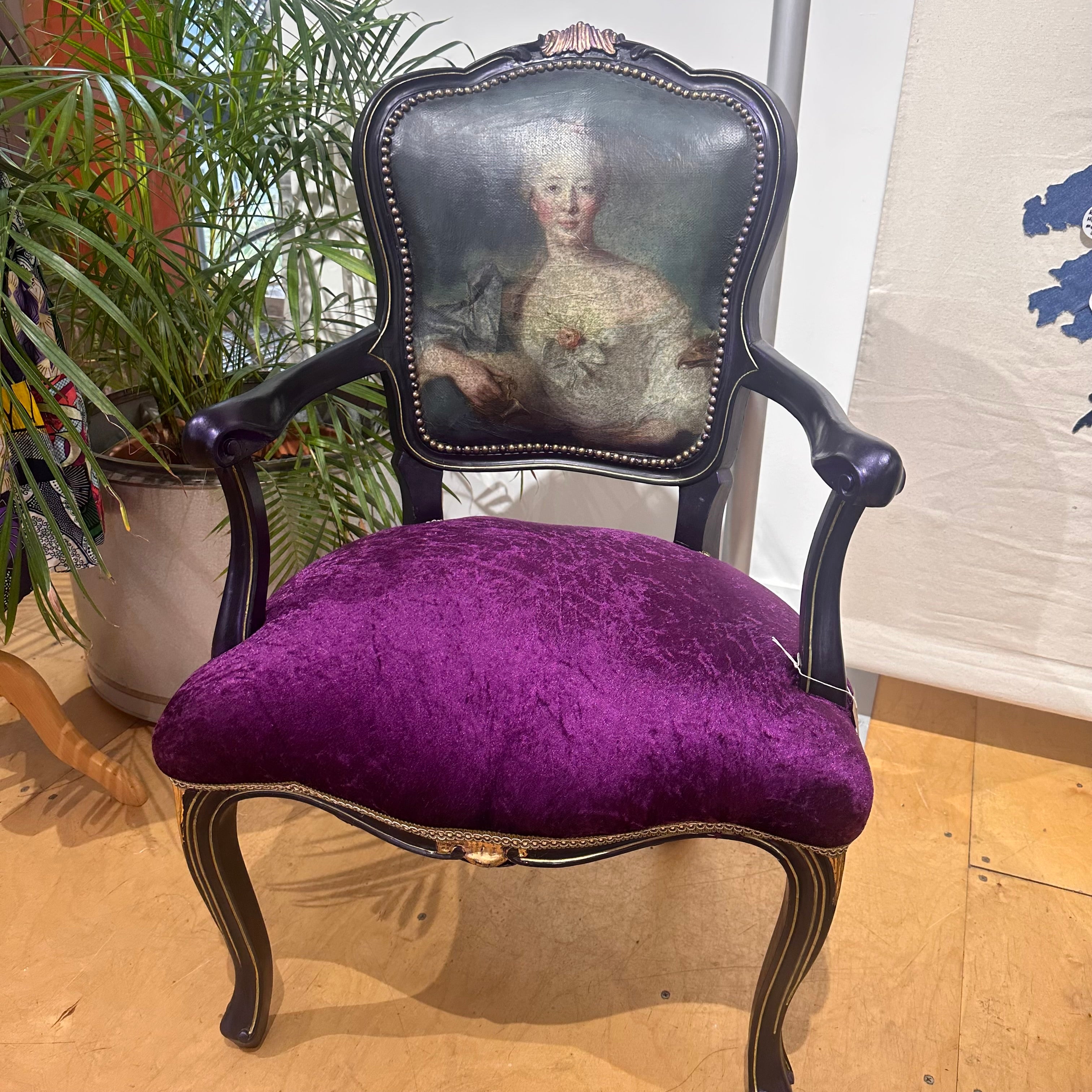 Purple upholstered chair with a decorative back featuring a portrait in a room with plants and a white wall.