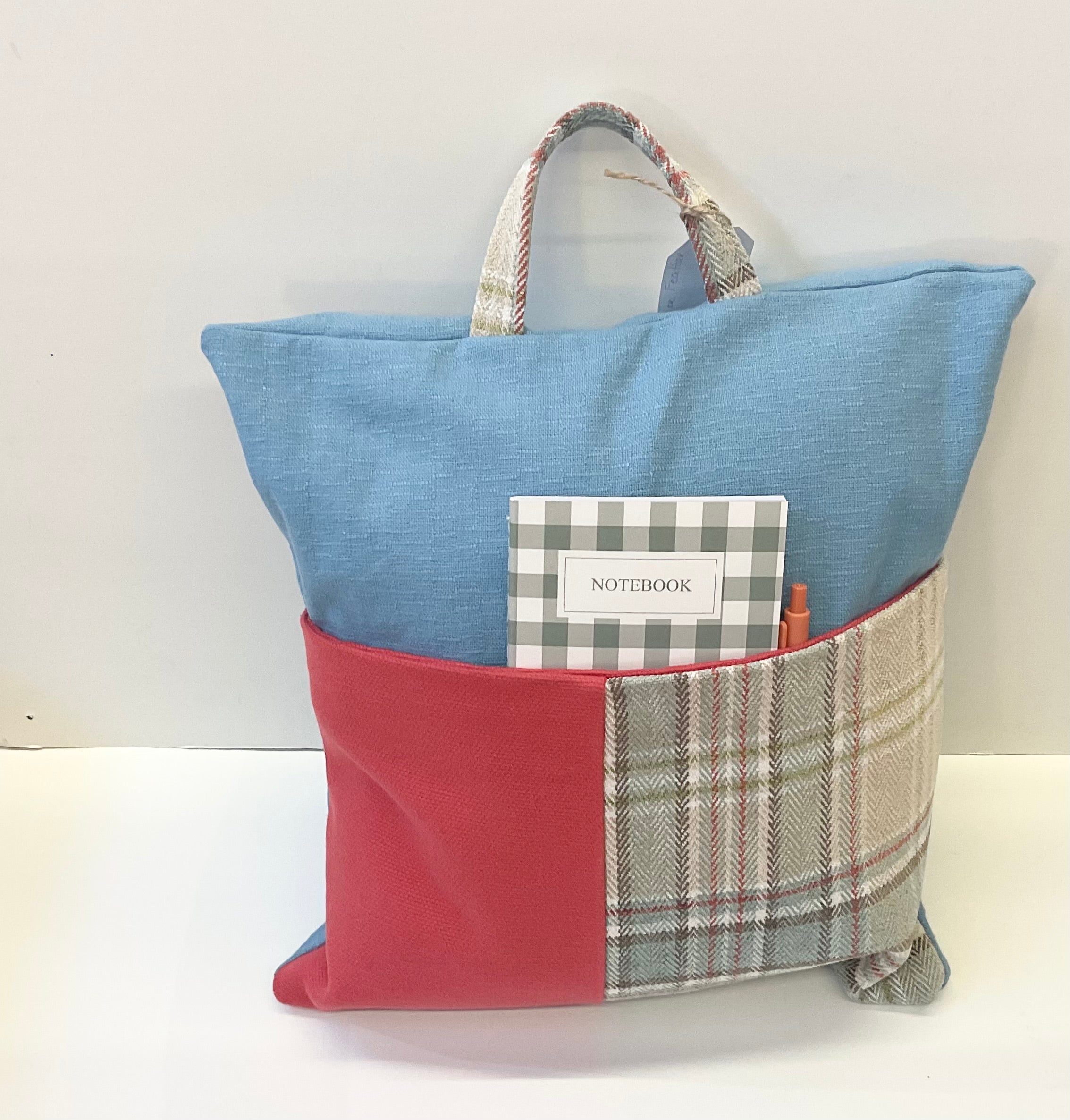 Blue pillow with a handle and a red and tweed pocket containing  a notebook and pen