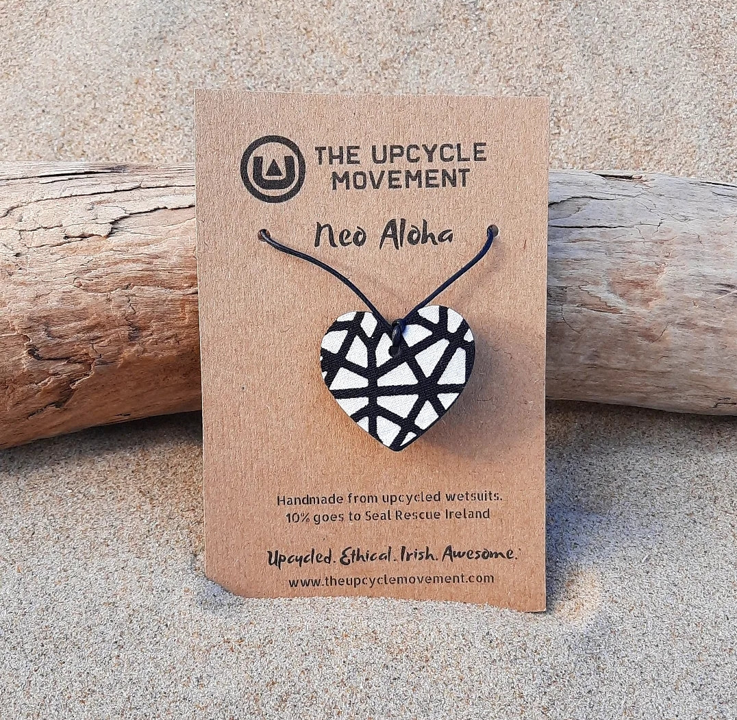 A necklace with a heart-shaped pendant featuring a black and white geometric pattern, displayed on a wooden surface with a brand tag attached.